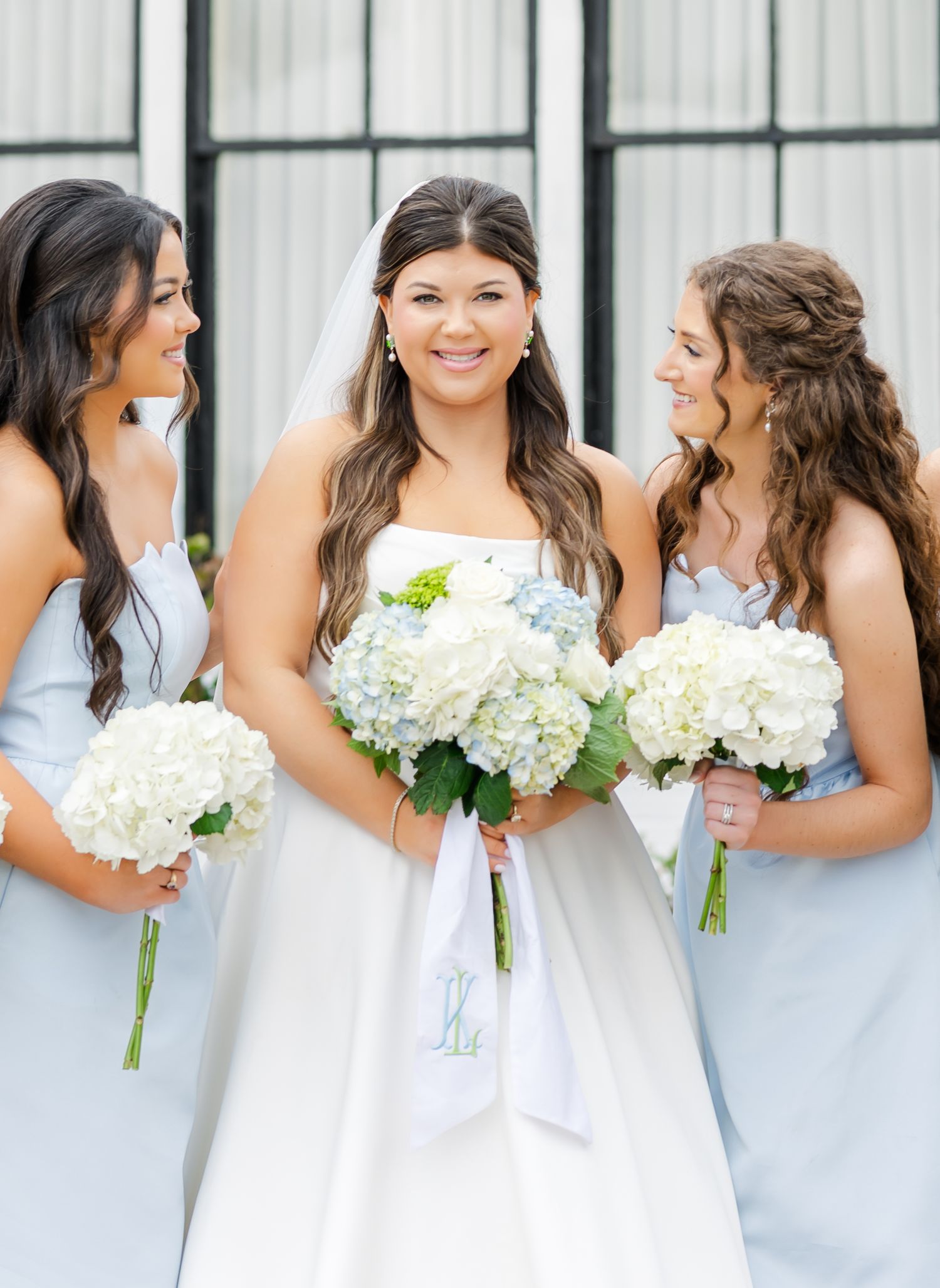 Smiling bride in white gown holds bouquet flanked by two bridesmaids in light blue dresses holding hydrangea bouquets.