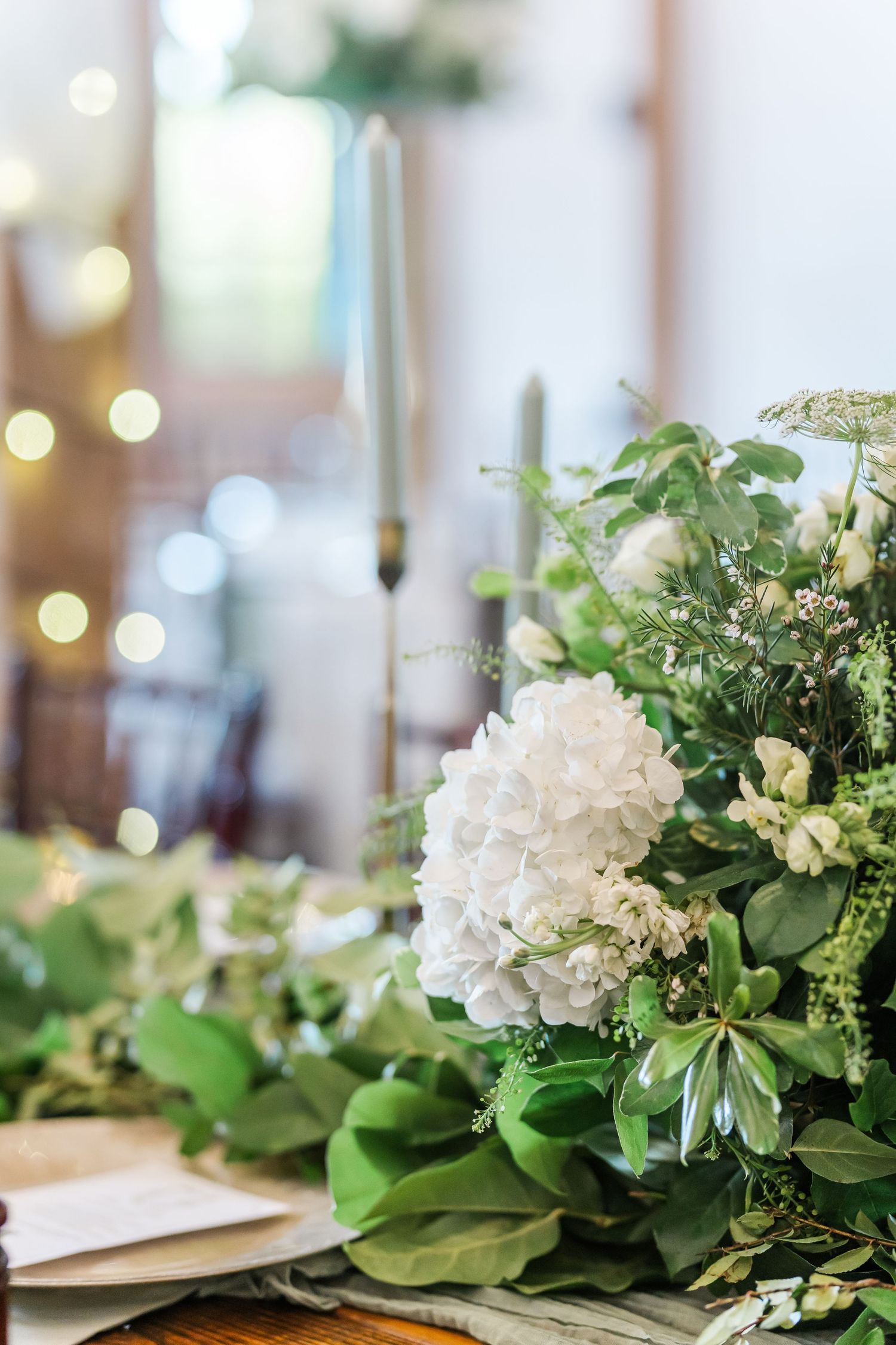Elegant white hydrangea and greenery centerpiece arrangements on a dining table with soft bokeh lighting in the background.