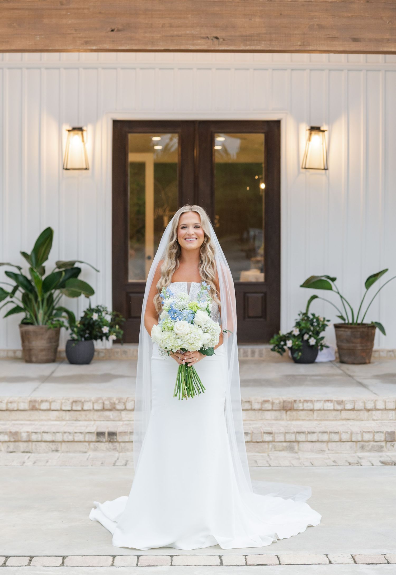 Radiant bride in strapless white gown with long veil holding blue and white bouquet in front of venue entrance.