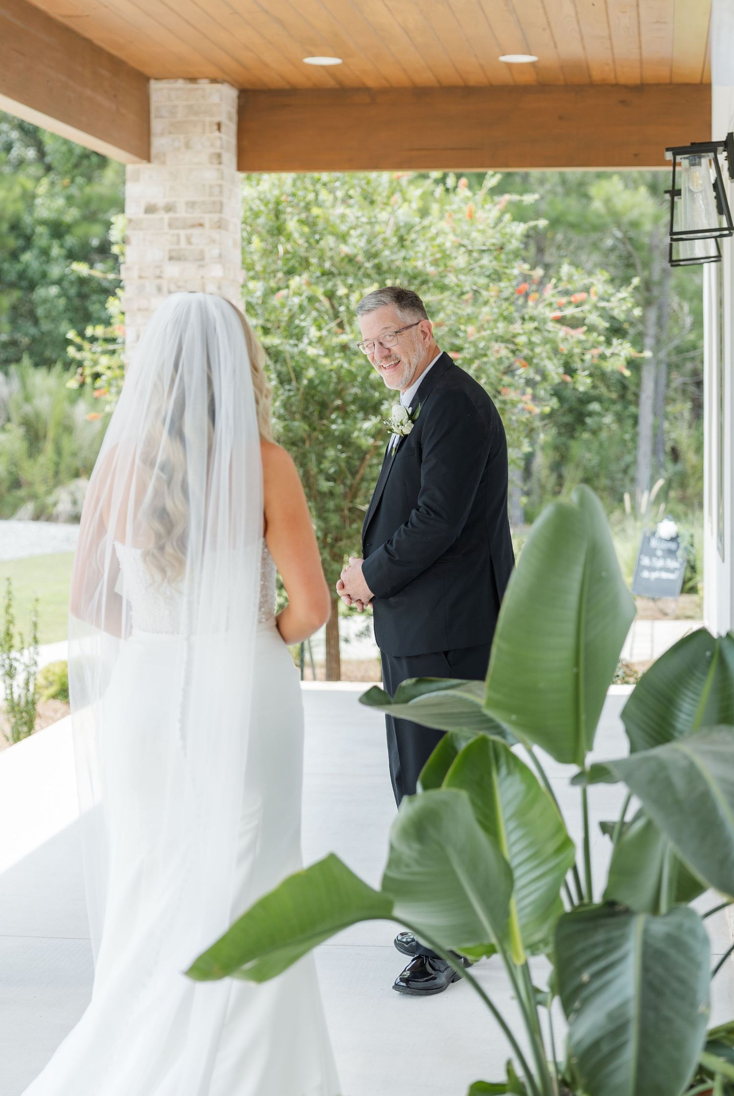 Bride in white gown and veil shares emotional first look moment with smiling father in black suit outdoors.