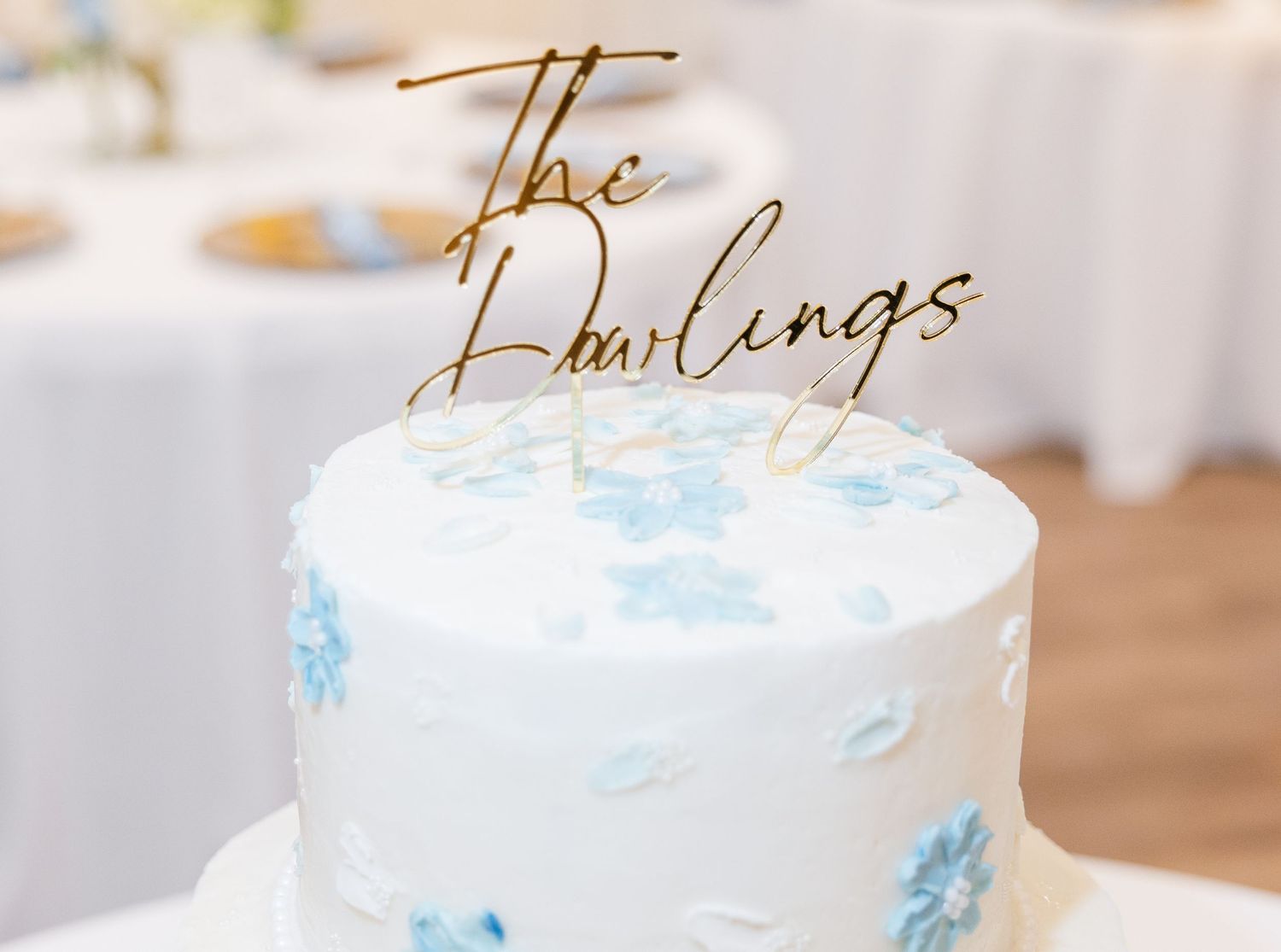 White wedding cake with blue floral details topped with gold mirror acrylic The Darlings cake topper.