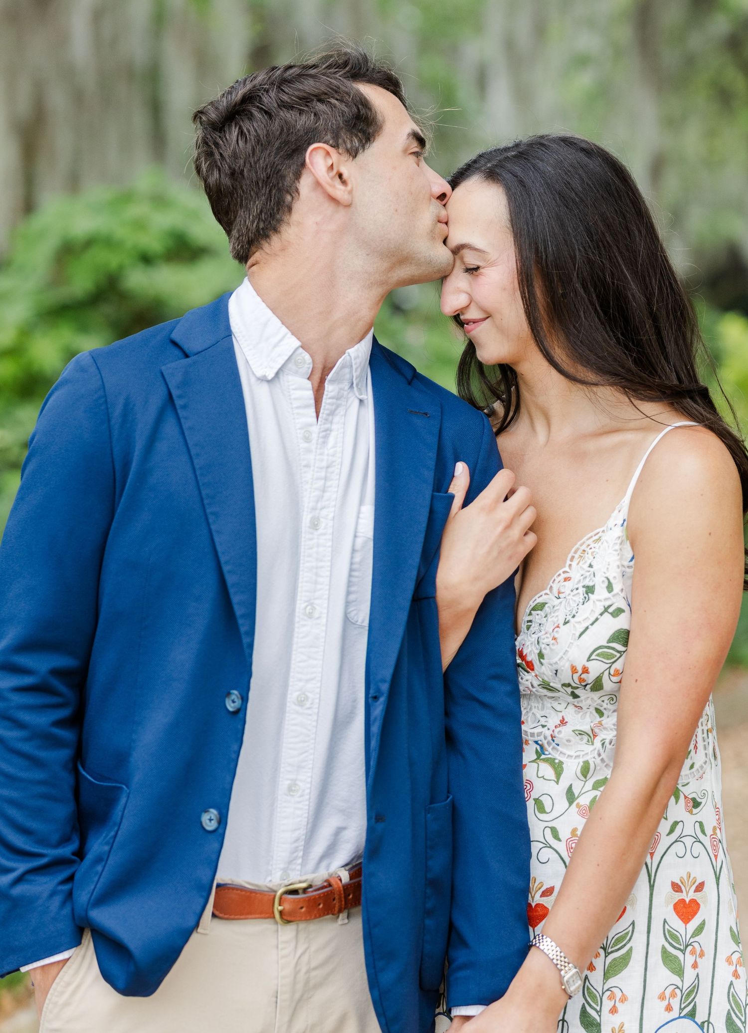 A couple shares a tender kiss outdoors, man in blue blazer and woman in floral dress, surrounded by lush greenery.