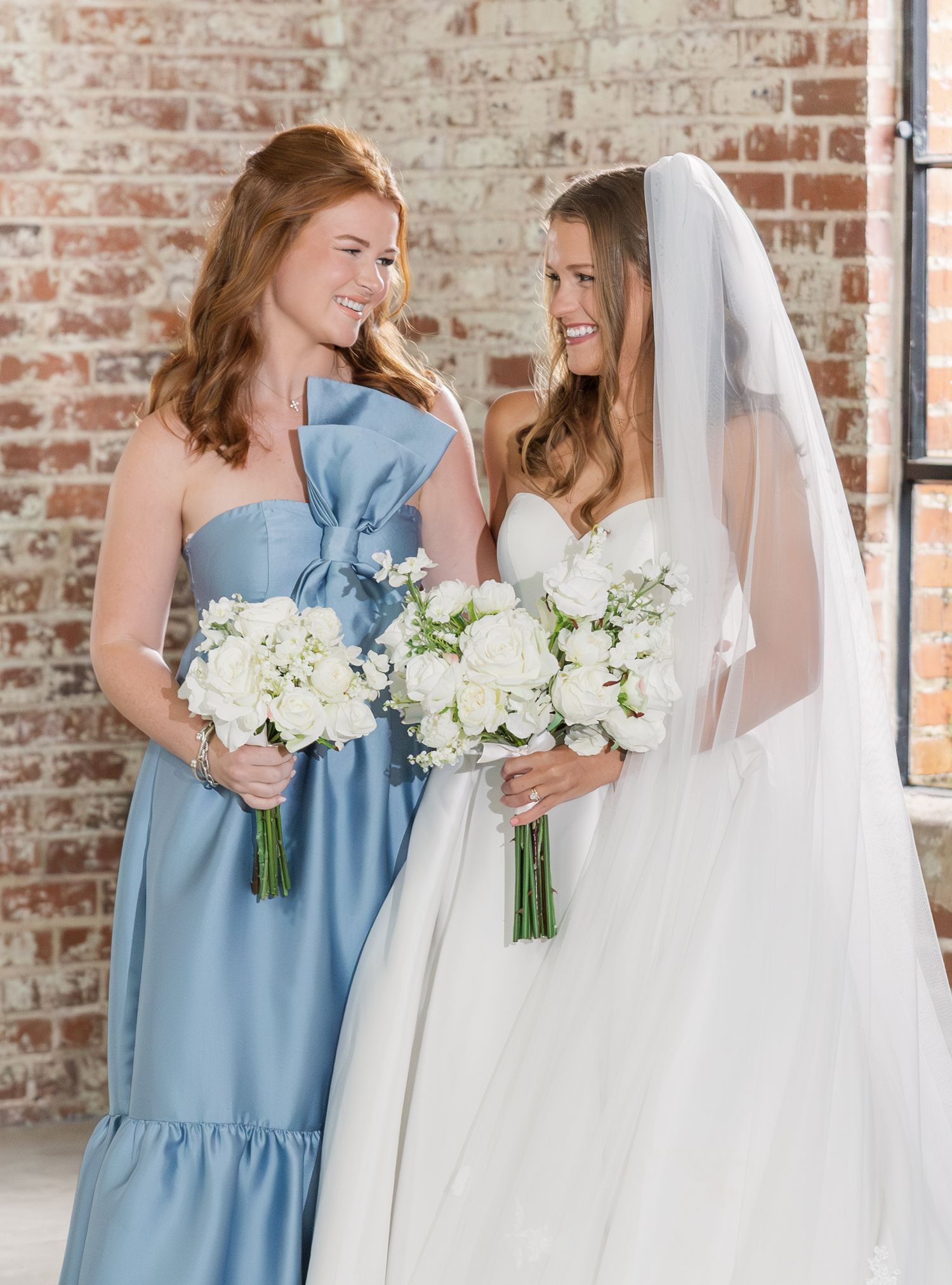 Bride in white gown and veil smiles with bridesmaid in blue dress, both holding white bouquets.