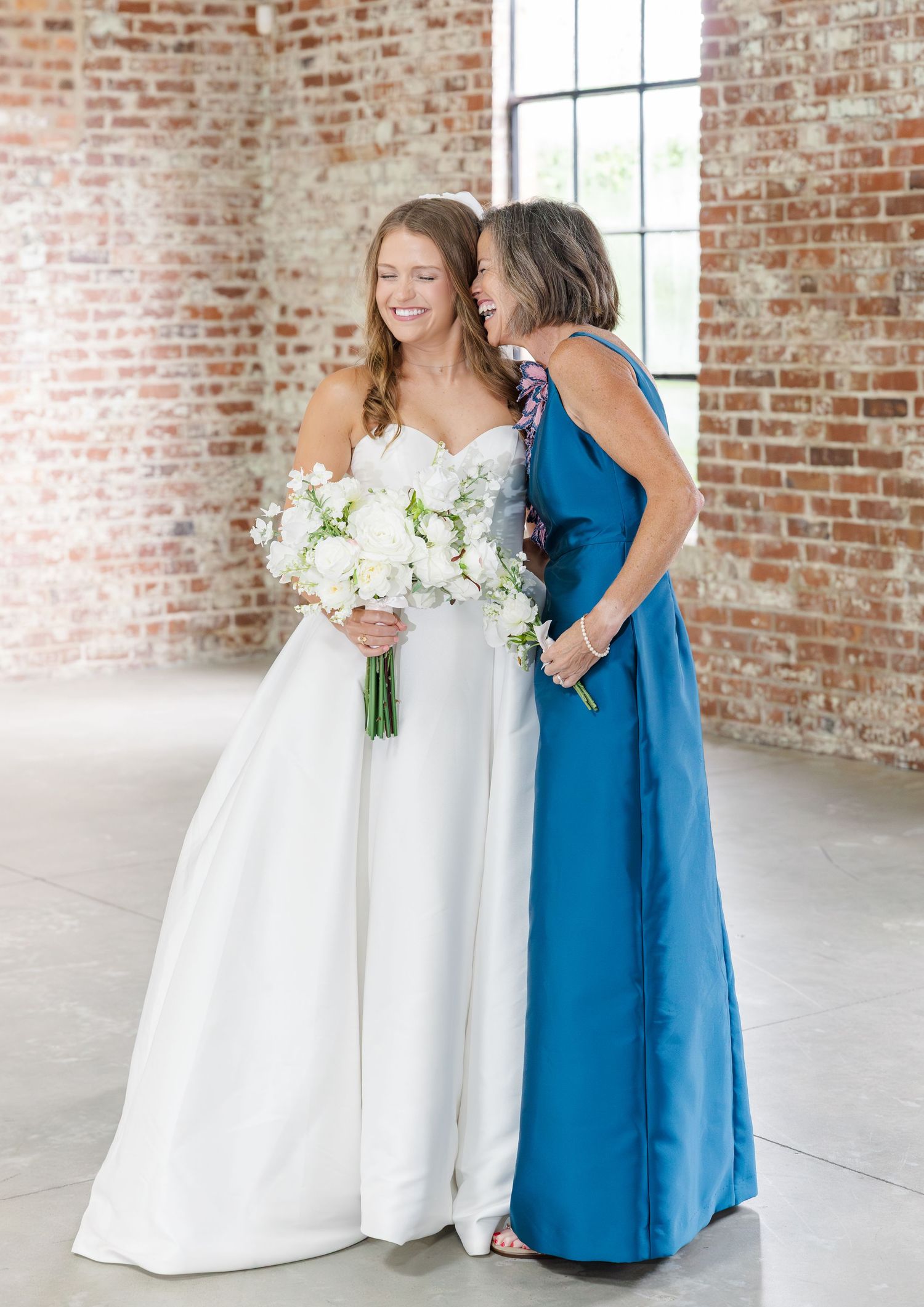 Bride in white gown holding bouquet smiles with woman in blue dress in brick venue.