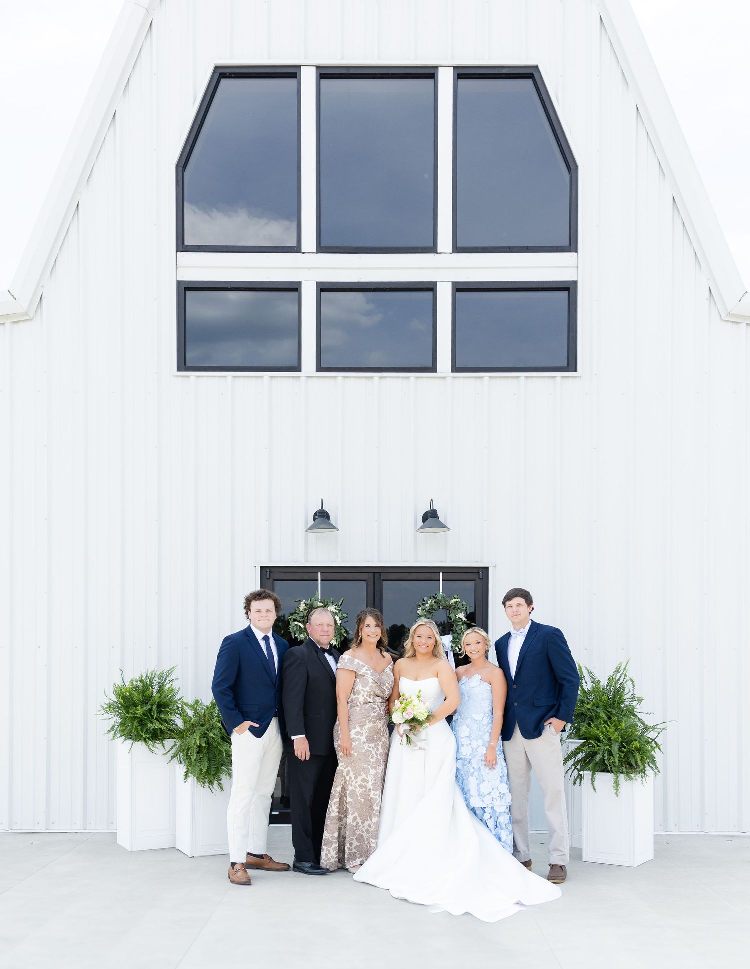 Wedding party poses outside a white barn venue with large windows, featuring bride in white gown with family in navy and floral attire.