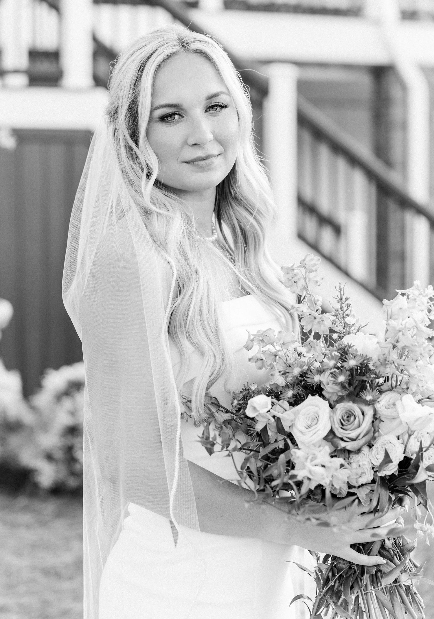 Black and white portrait of blonde bride in veil holding lush floral bouquet outdoors.
