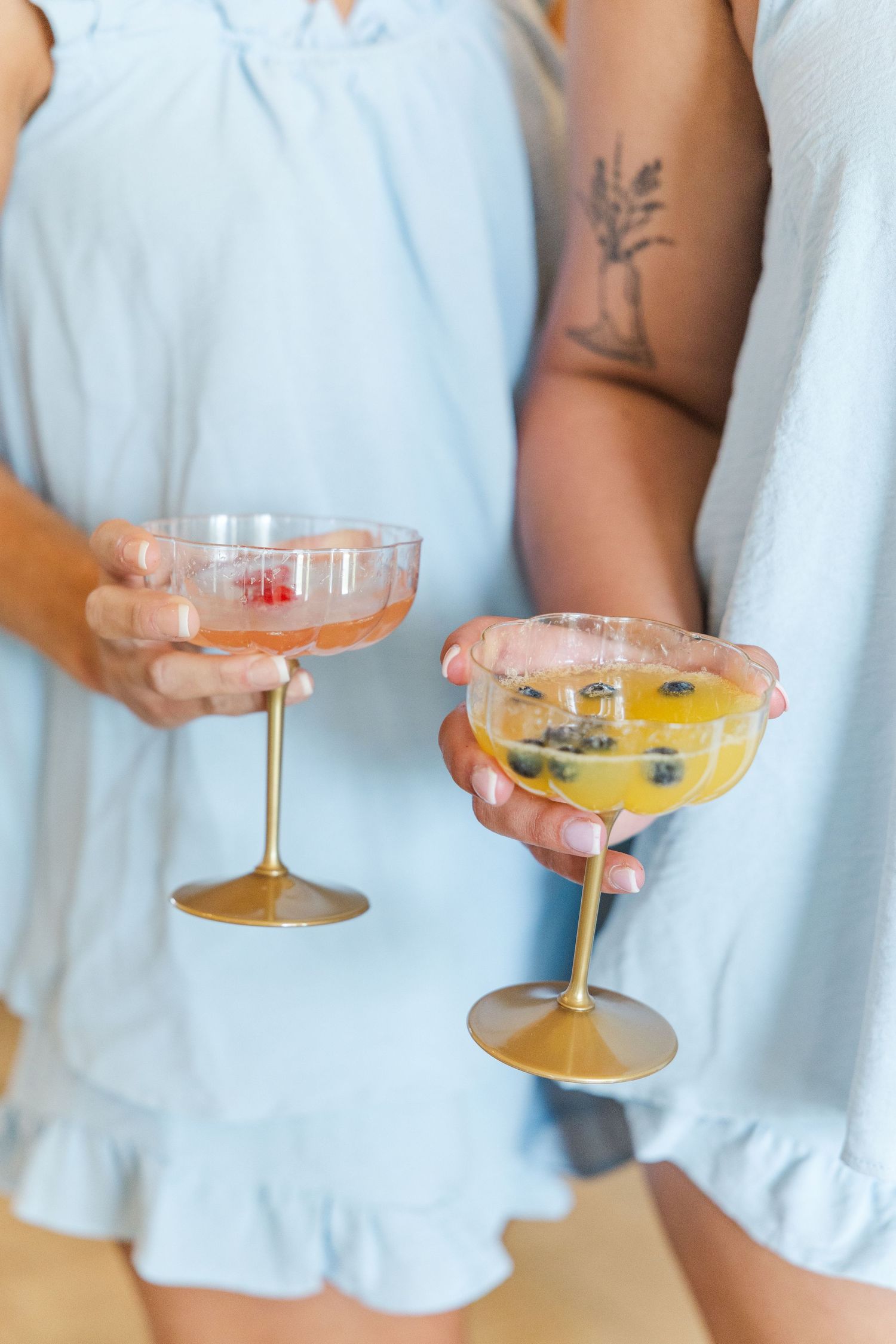 Two women in blue dresses holding gold-stemmed coupe cocktail glasses with colorful fruit garnishes.