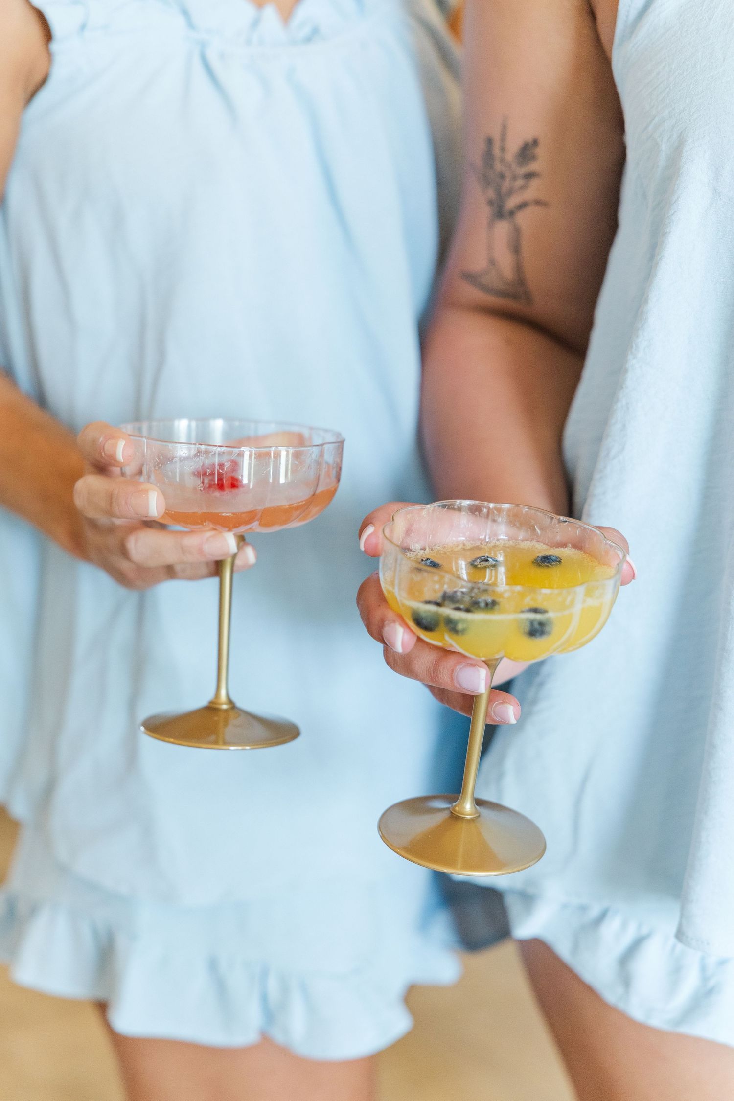 Two women in light blue dresses holding elegant gold-stemmed coupe cocktail glasses with colorful drinks.