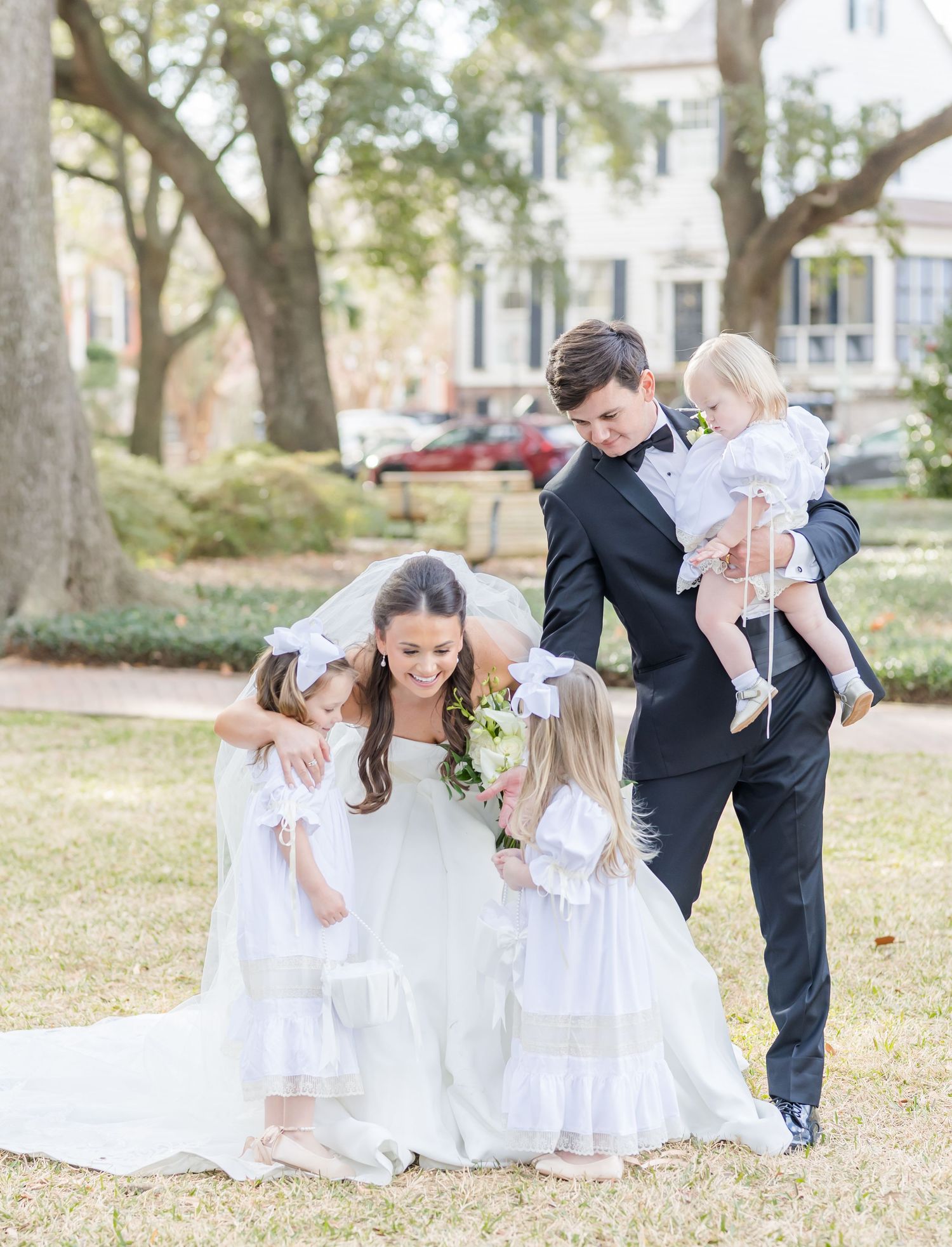 Bride and groom with flower girls in white dresses celebrating outdoors under oak trees.