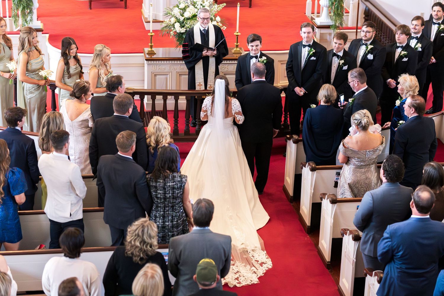 Bride walks down church aisle in white gown with guests seated in wooden pews during traditional wedding ceremony.