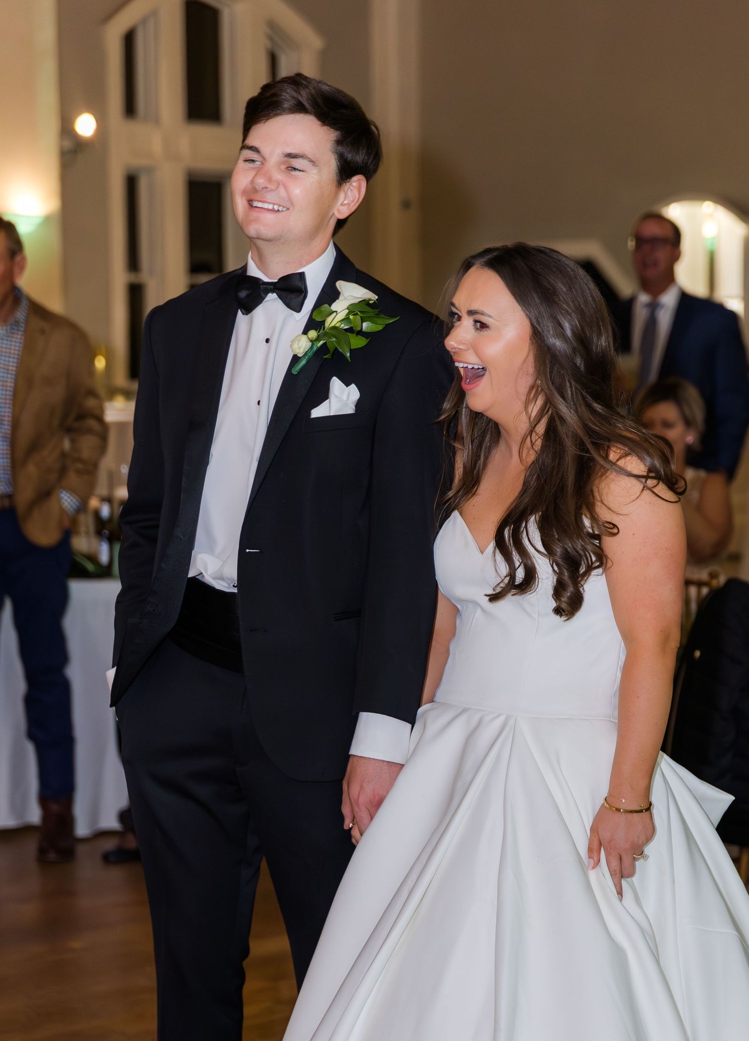 Bride and groom smiling joyfully at their wedding reception, groom in black tuxedo, bride in white gown.