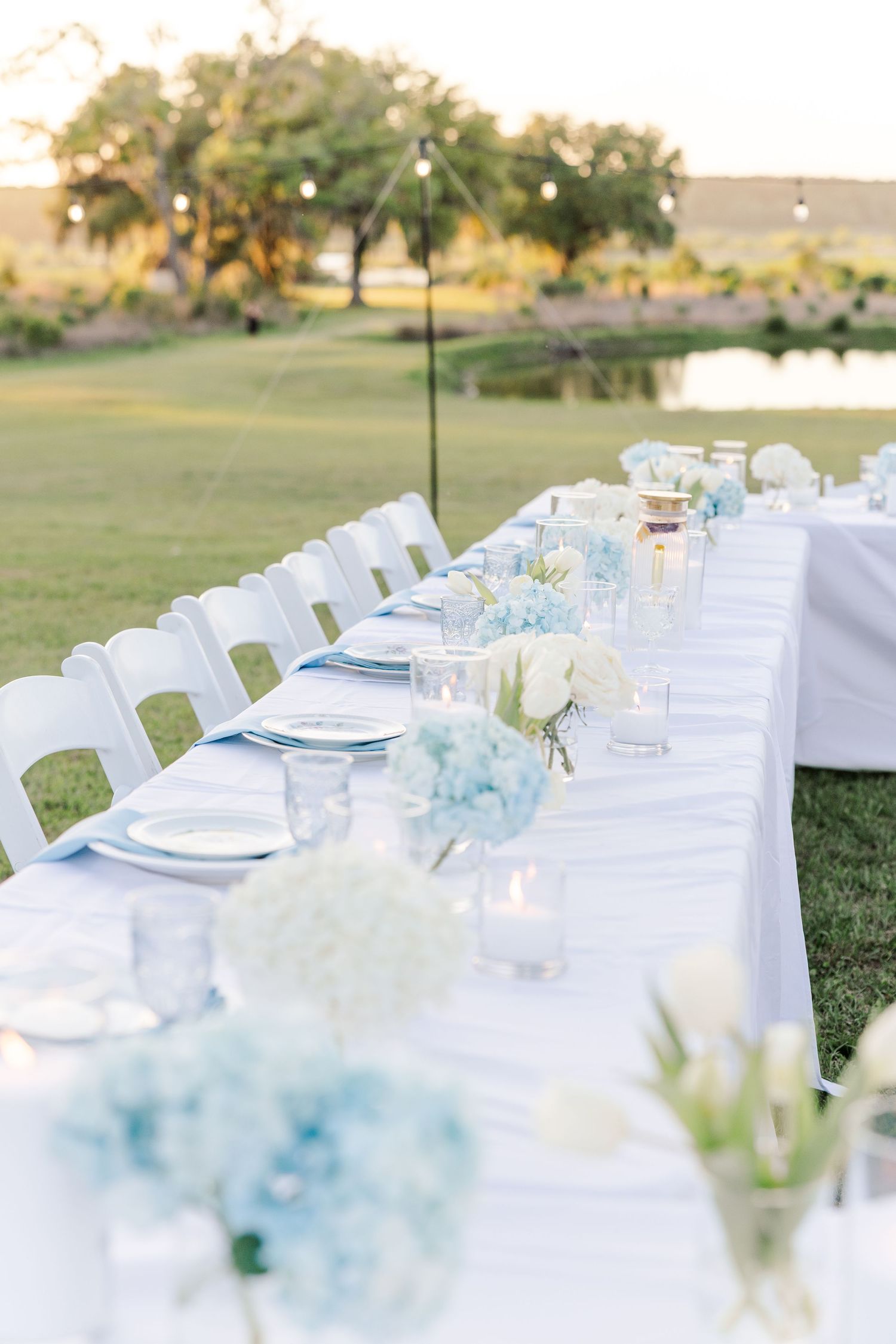 Outdoor wedding reception long table with white linens, light blue hydrangeas, candles, and white chairs at golden hour.