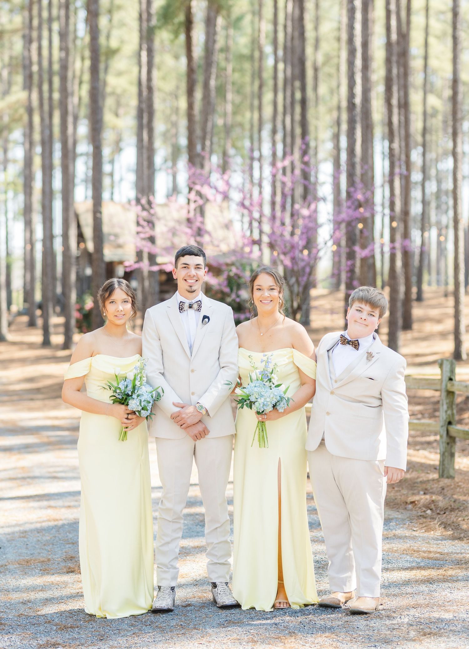 Wedding party in yellow bridesmaid gowns and tan suits posing among pine trees with spring blooms.