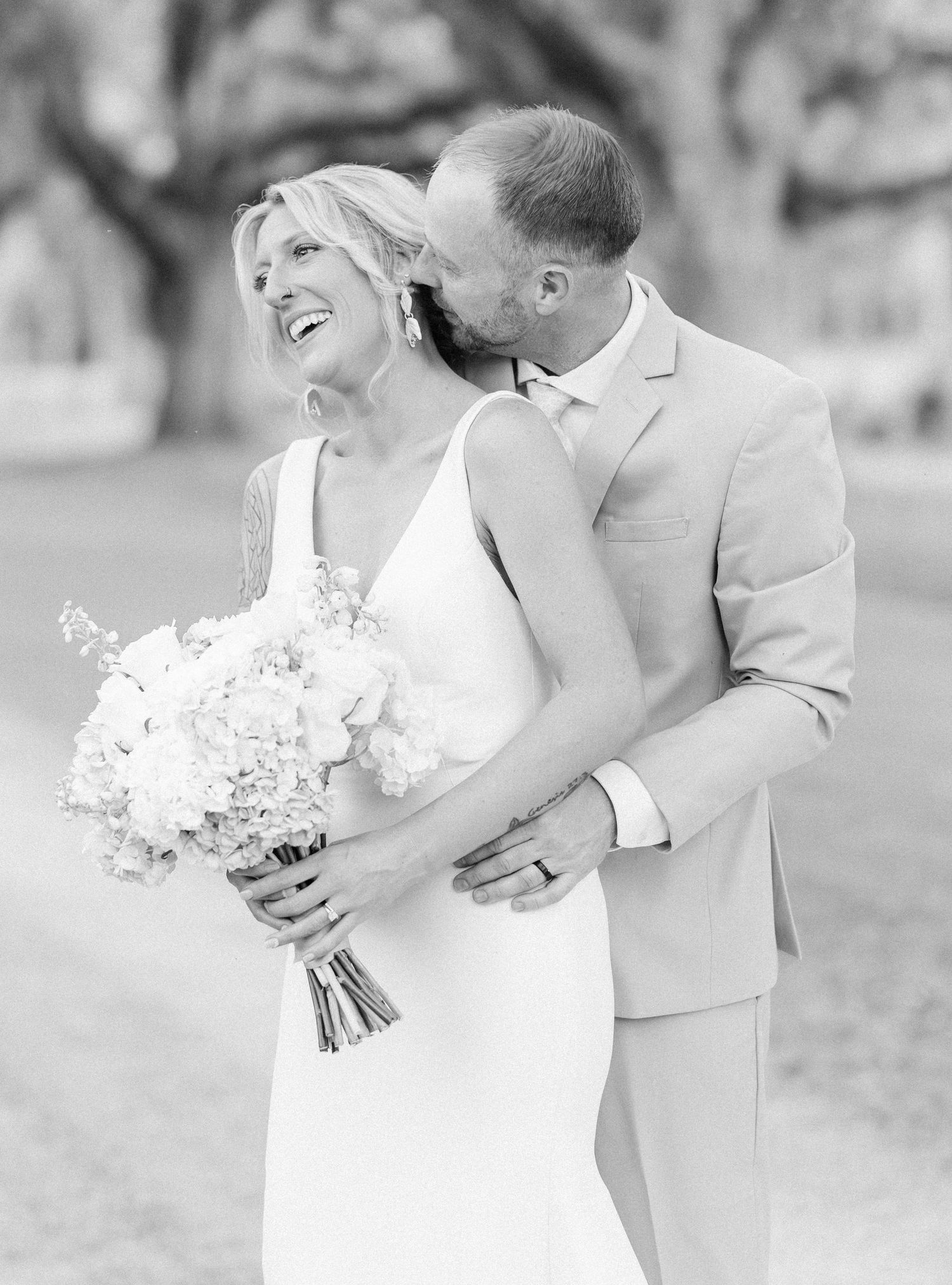 Black and white photo of laughing bride holding bouquet while groom kisses her cheek at outdoor wedding.