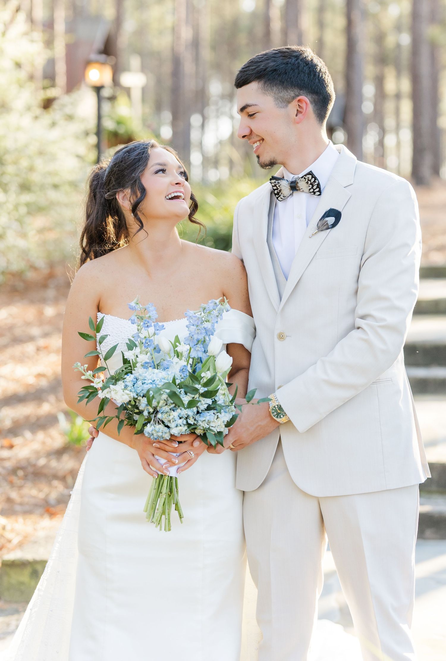 Smiling bride holding blue floral bouquet stands with groom in tan suit and leopard bow tie outdoors.