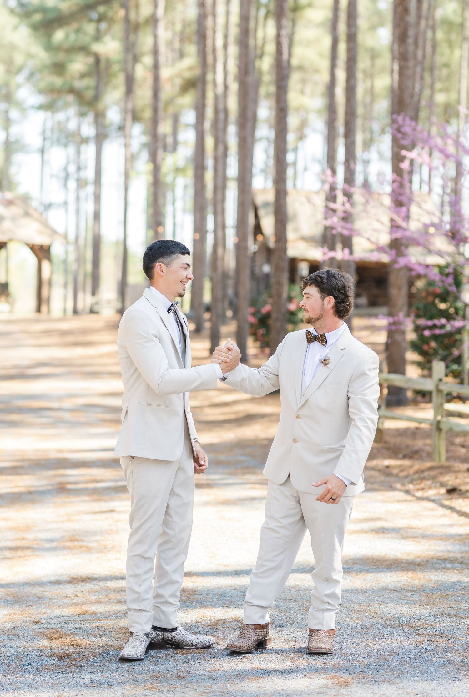 Two groomsmen in beige suits shaking hands on a gravel path with blooming trees behind.