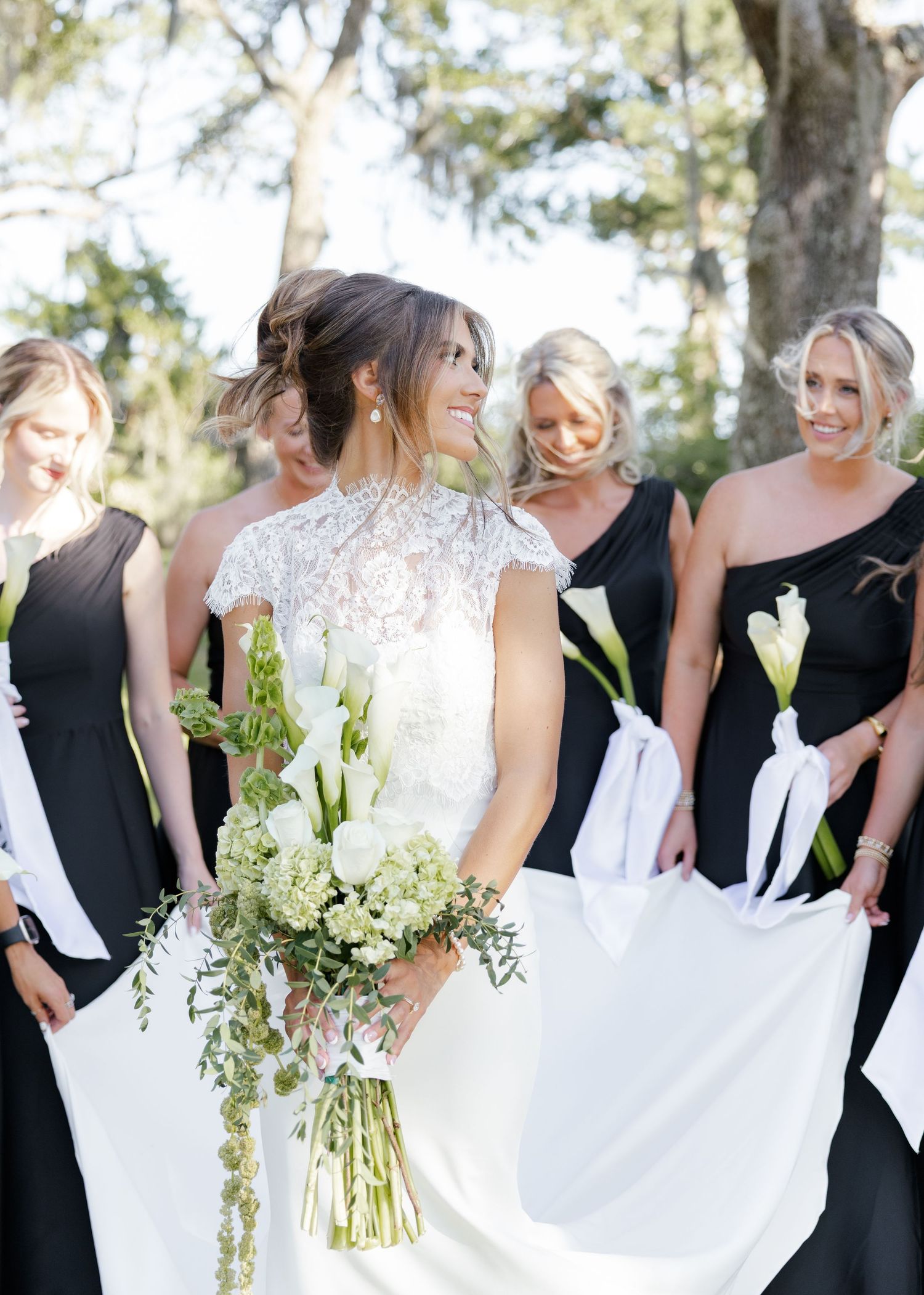 Bride in lace back wedding dress laughs with bridesmaids in black gowns holding white calla lilies outside.