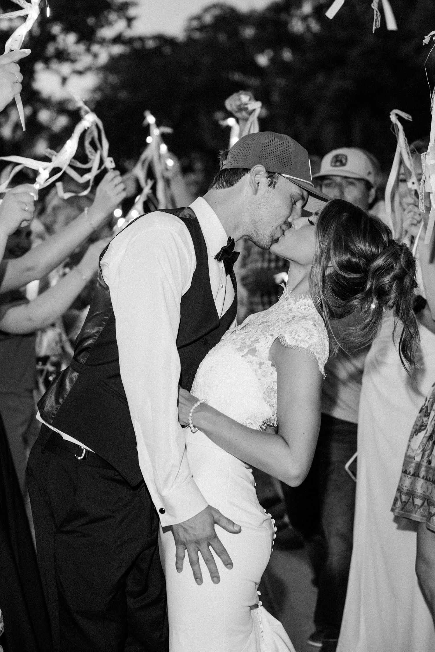 Black and white photo of a bride and groom sharing a kiss during a ribbon wand sendoff at their wedding reception.