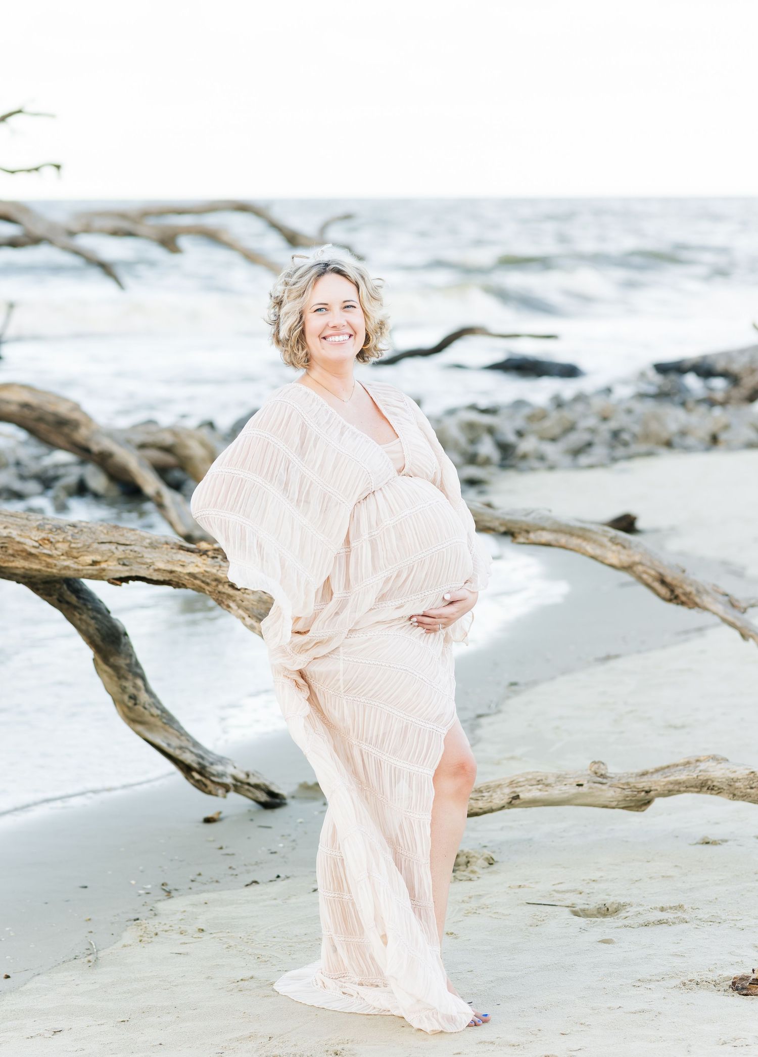 Ethereal maternity photography session on a driftwood-strewn beach with flowing peach dress catching the coastal breeze.