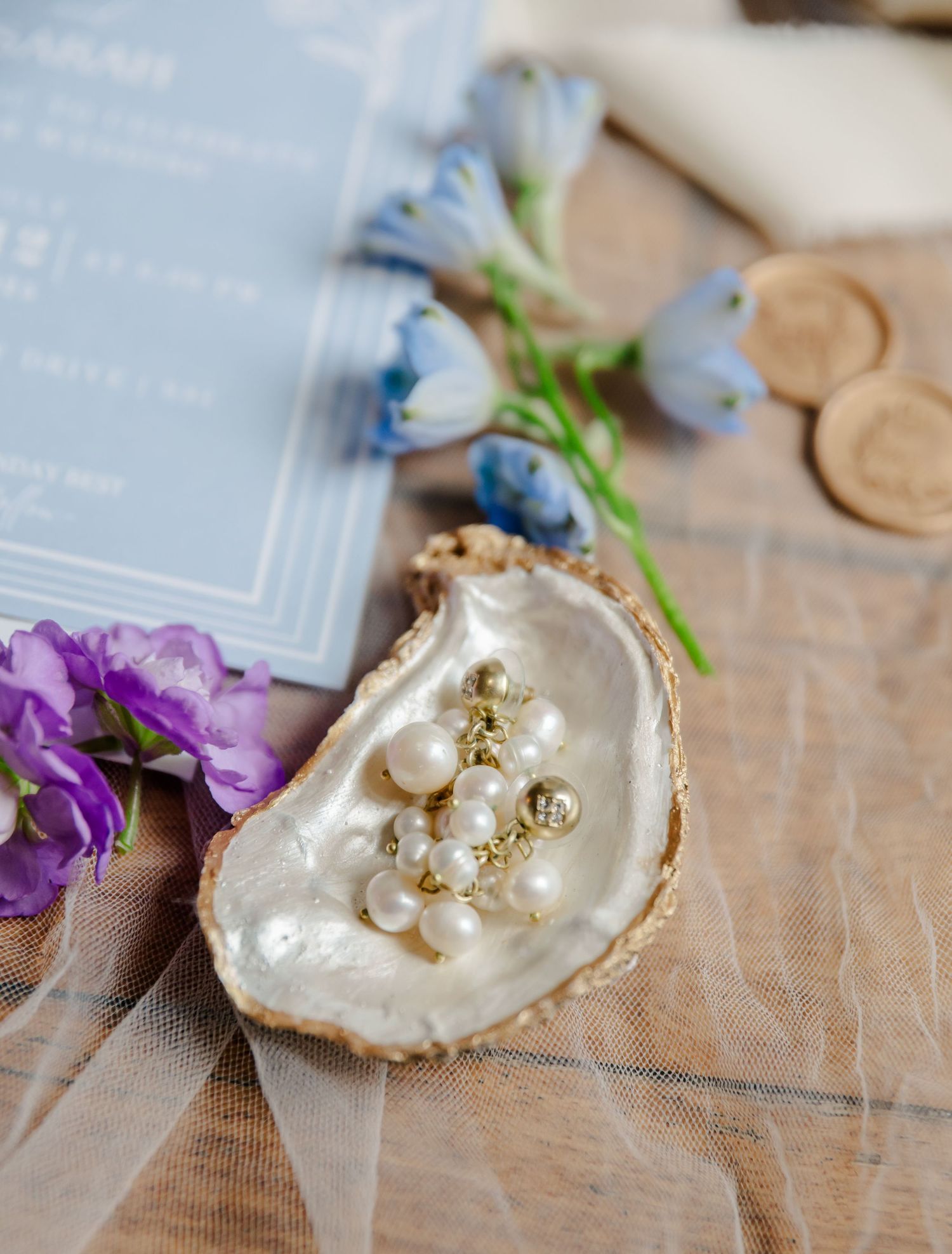 A pearlescent oyster shell ring dish with gold embellishments and delicate purple flowers beside it on a linen surface.