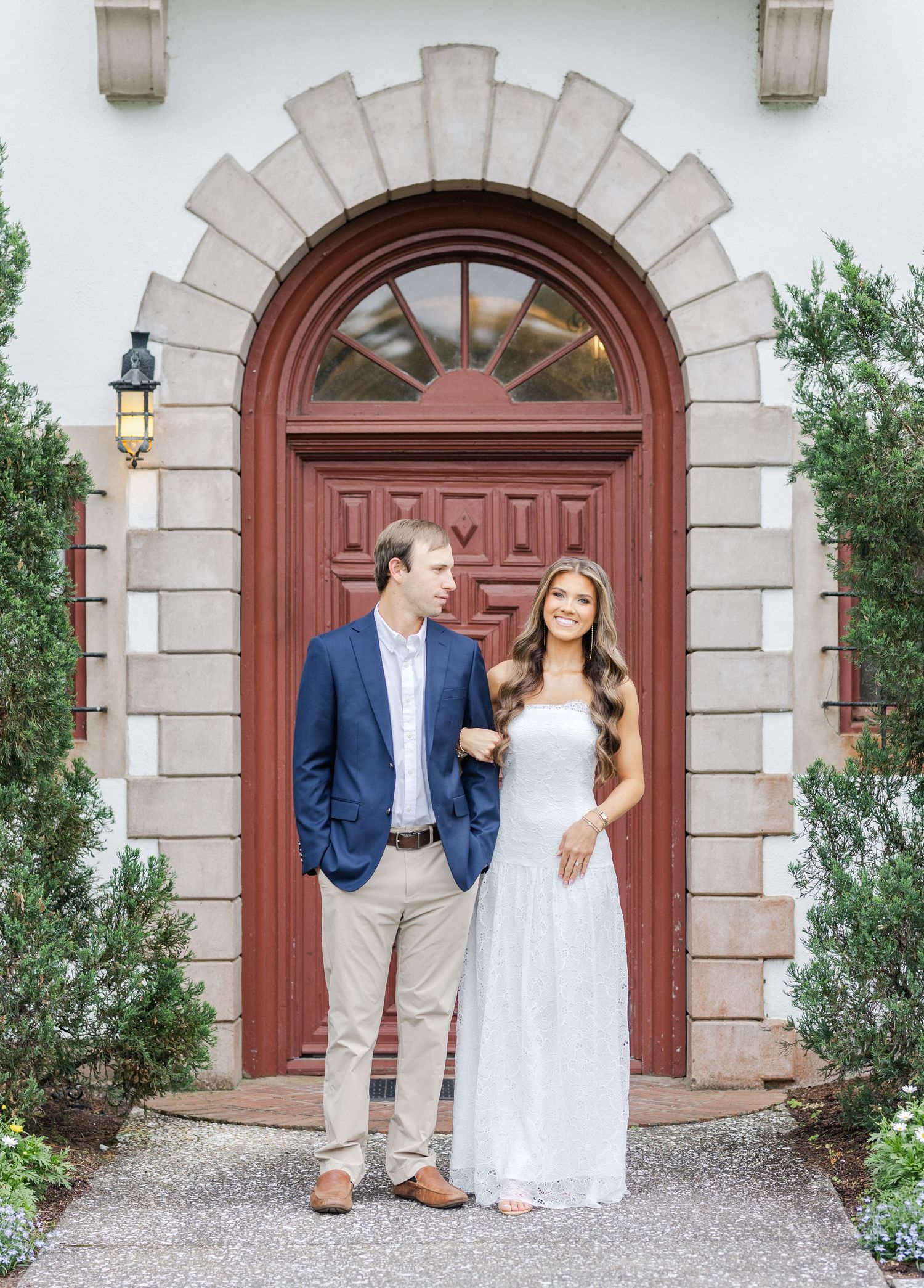 Couple posing in front of elegant arched red door, man in navy blazer and woman in white dress, engagement photo.