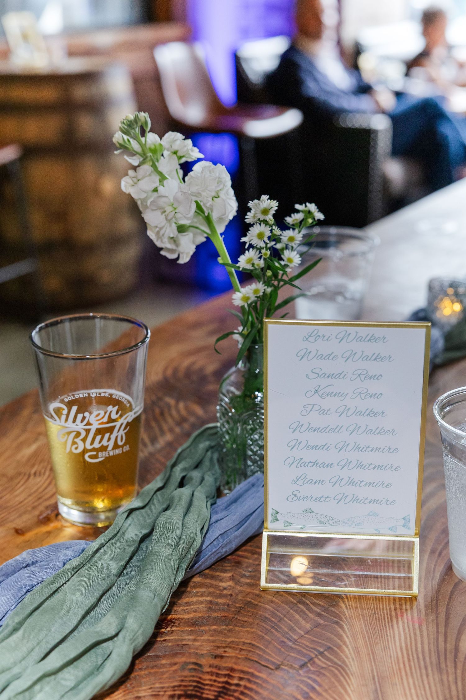 Rustic wedding reception table with River Bluff beer glass, white floral arrangement, and elegant seating chart card.