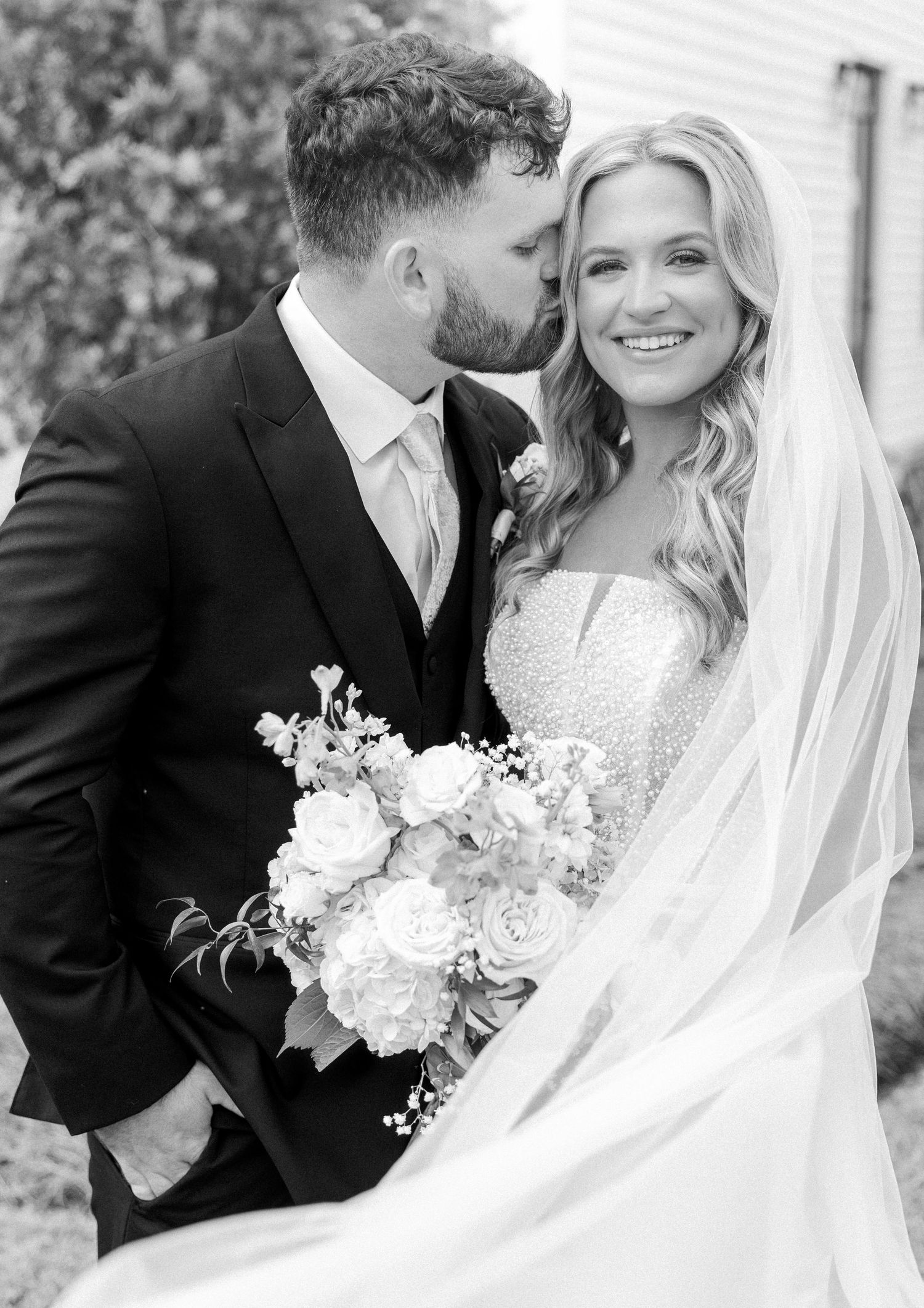 Black and white photo of groom kissing bride on cheek as she smiles holding a lush white rose bouquet.