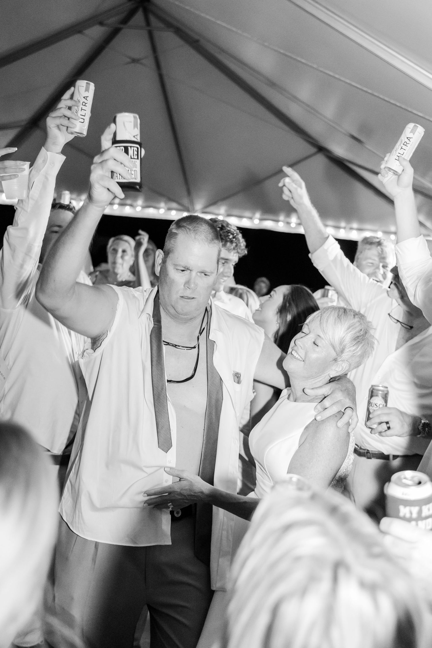 Crowd of wedding guests dancing and celebrating with raised arms under a white tent.