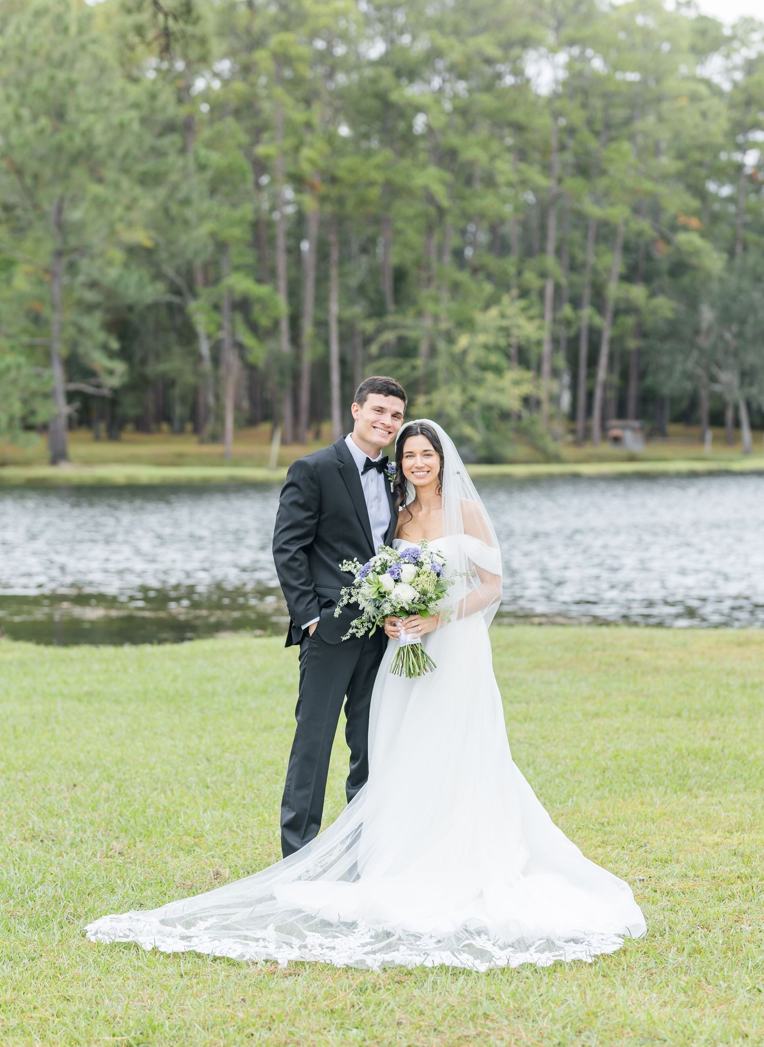 A romantic wedding couple poses together on a grassy lawn beside a lake with trees in the background.