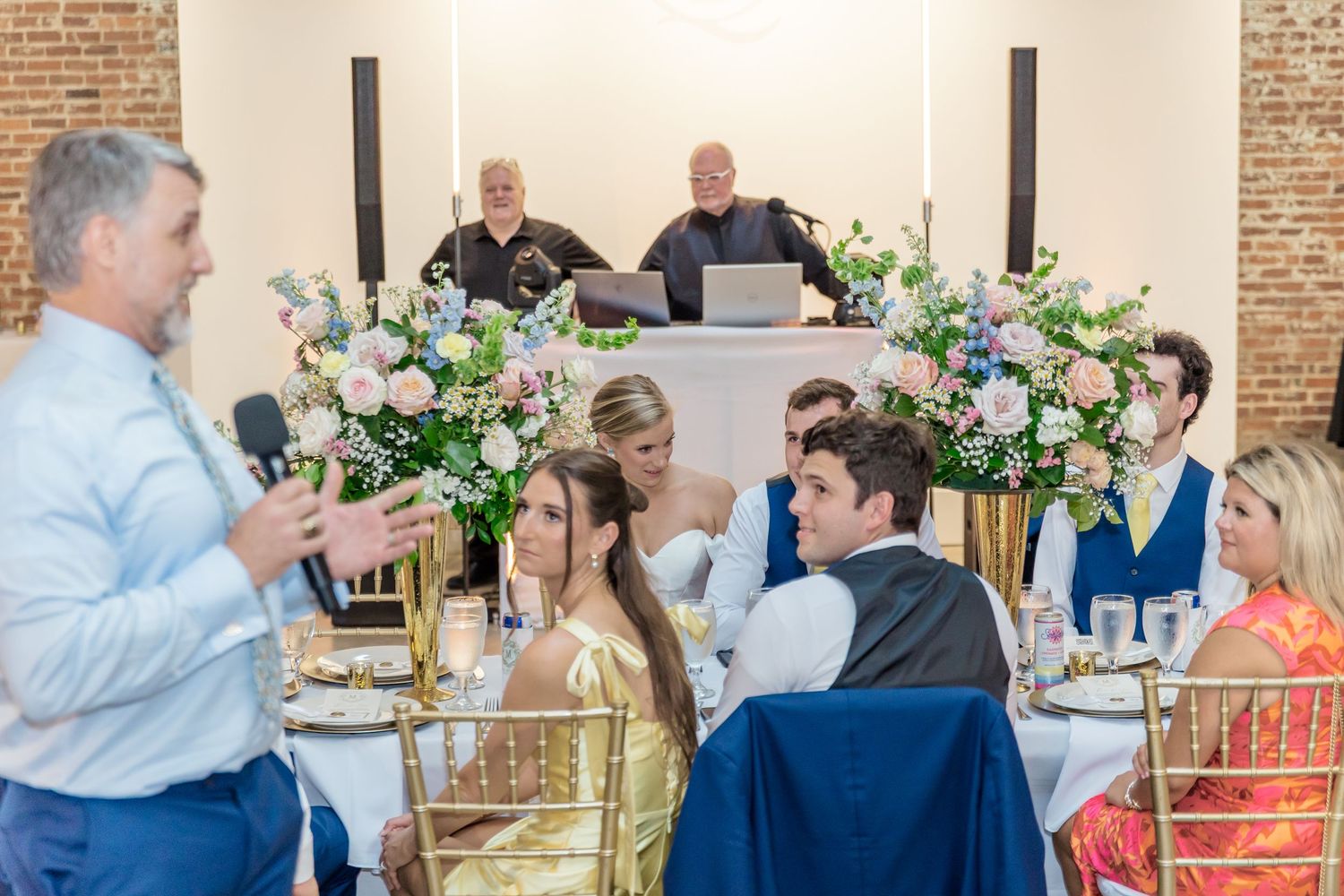 Wedding guests mingle and socialize during an elegant reception in a rustic brick-walled venue with floral arrangements.