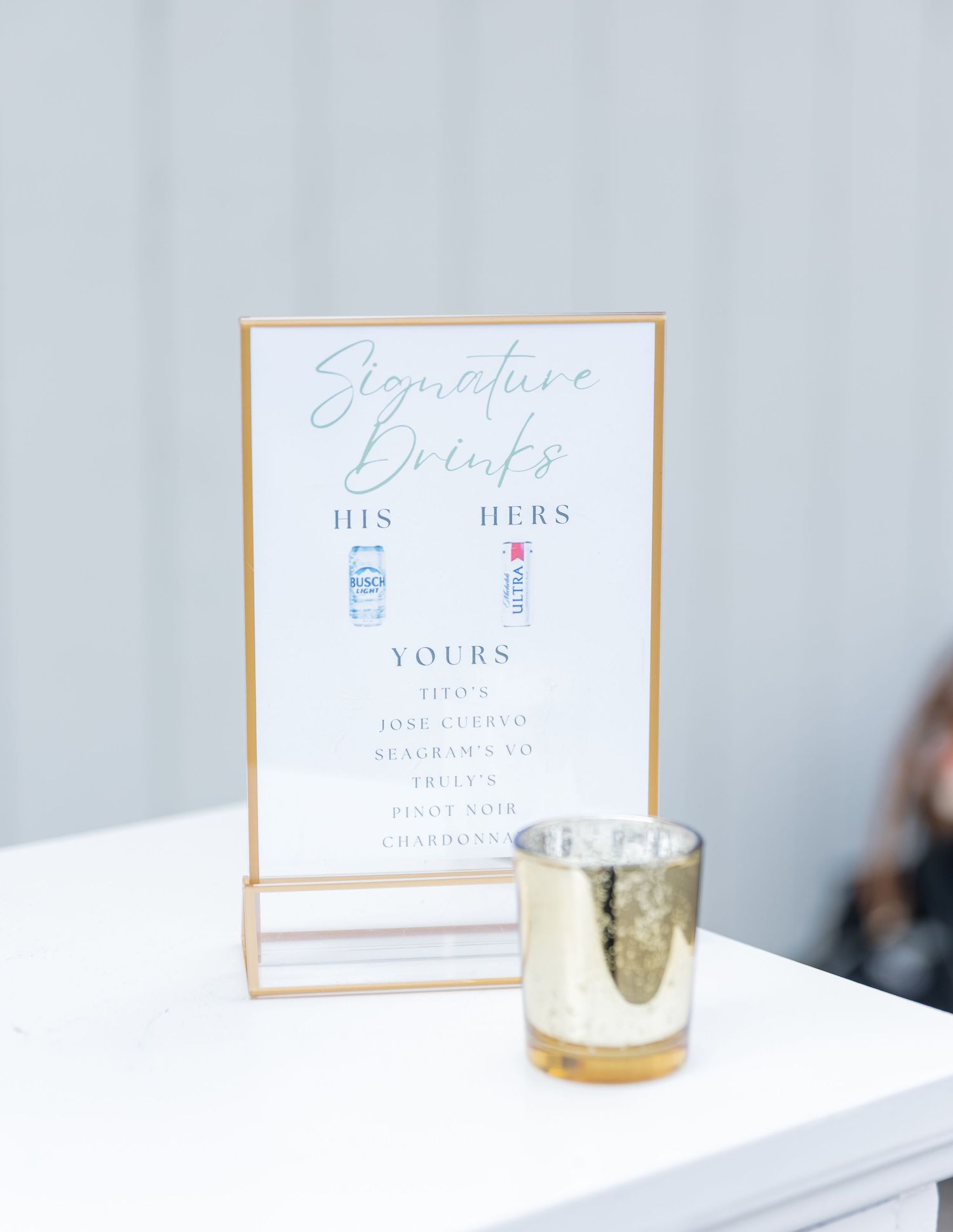 Elegant white and gold wedding signage displays in calligraphy against a light gray background with a votive candle.