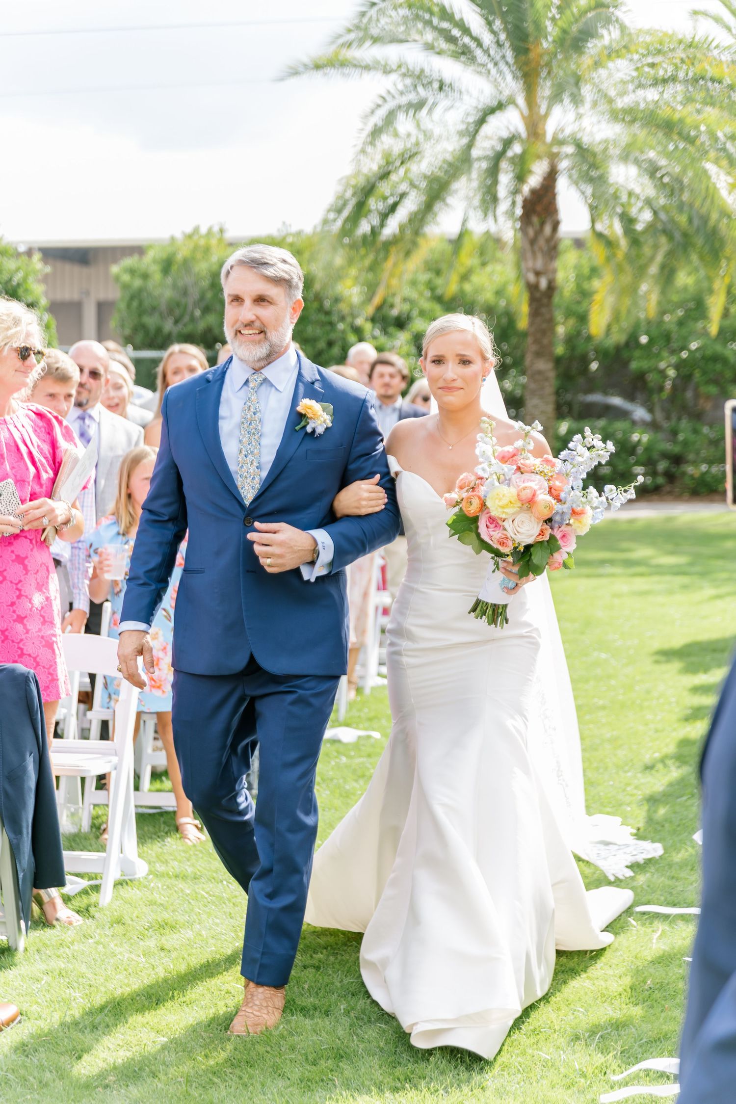 Wedding party celebrates on a sunny lawn with palm trees as guests look on during an outdoor ceremony.