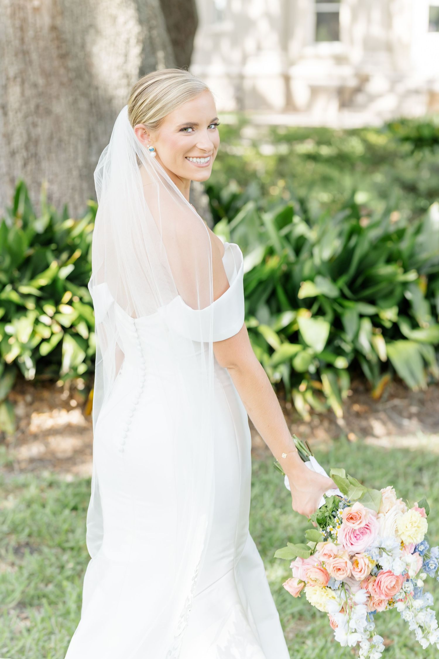 Bride in elegant white dress holds colorful bouquet of pink and blue flowers in outdoor garden setting.