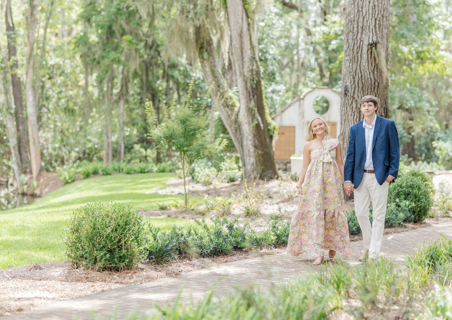 A couple strolls along a garden path surrounded by Spanish moss and lush greenery.