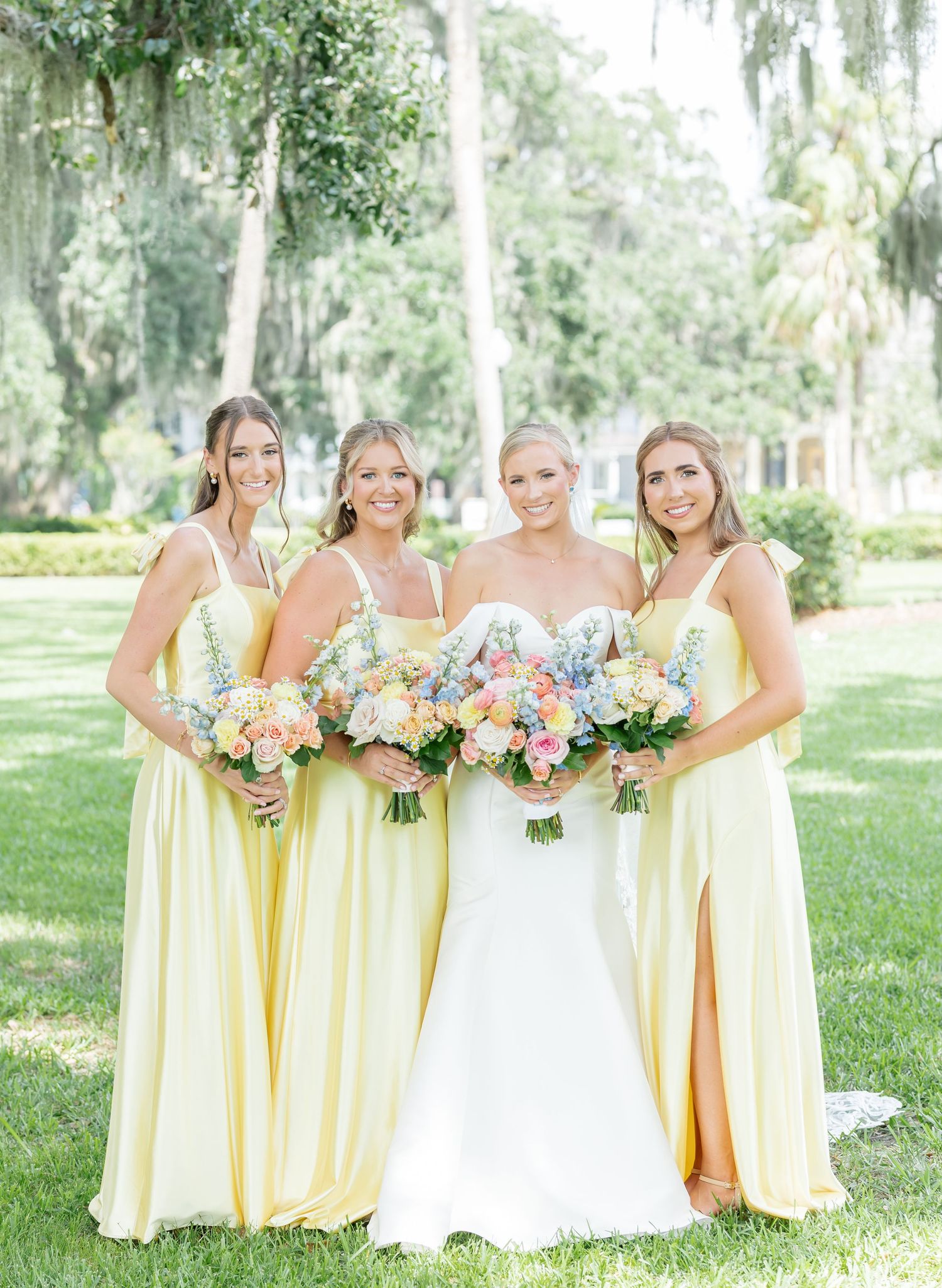 Bridal party in pale yellow bridesmaid dresses and white wedding gown pose together in a sunny garden setting.