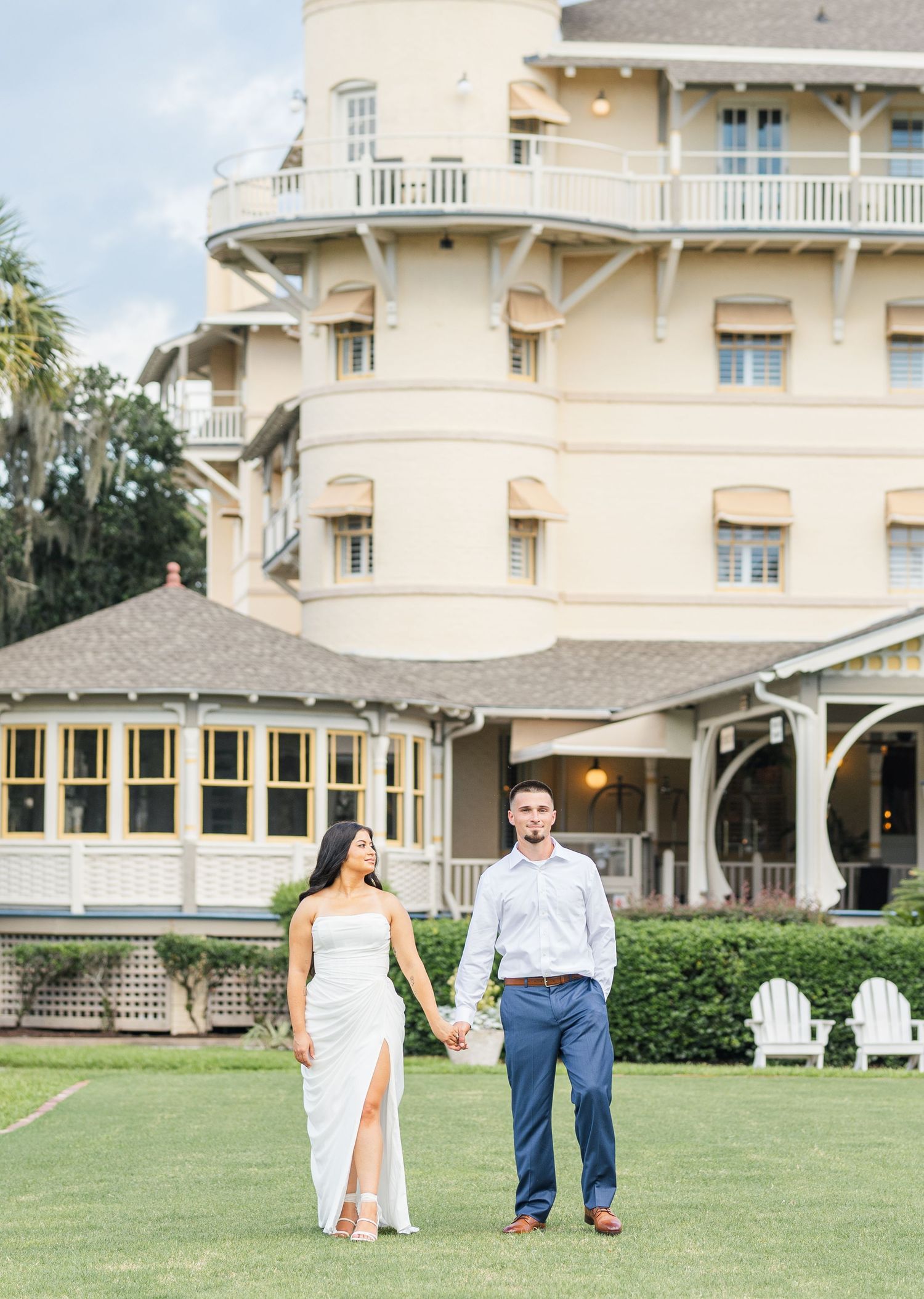 Couple holding hands in front of elegant yellow historic building on green lawn, woman in white dress, man in blue pants.