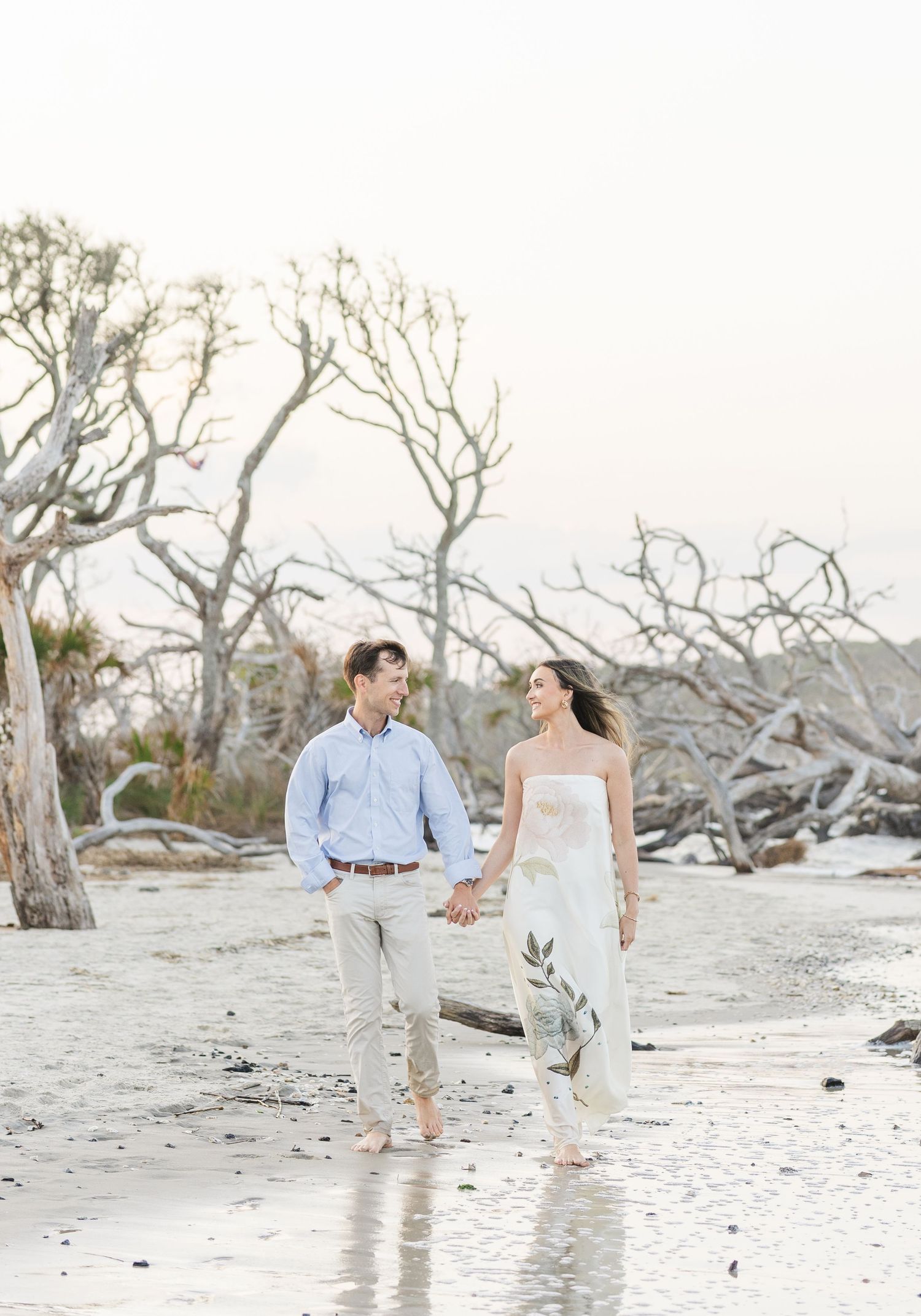 Couple strolling barefoot on beach holding hands, surrounded by driftwood trees in soft golden light.