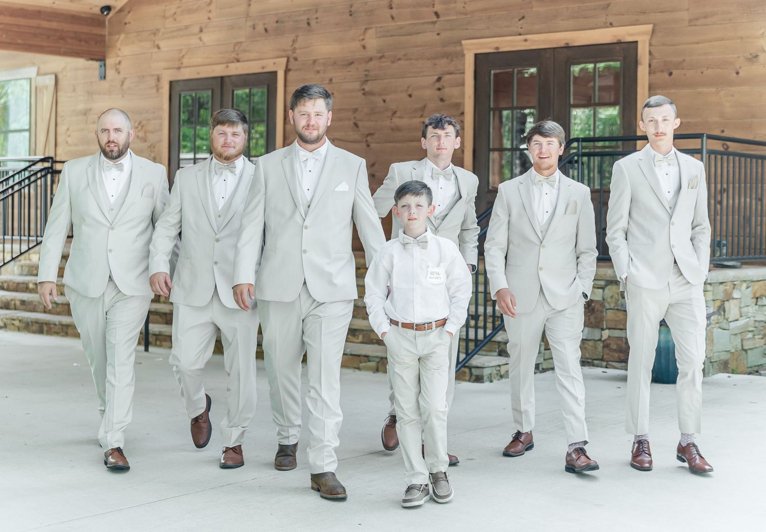 Wedding party in coordinated beige suits walks together on rustic porch.