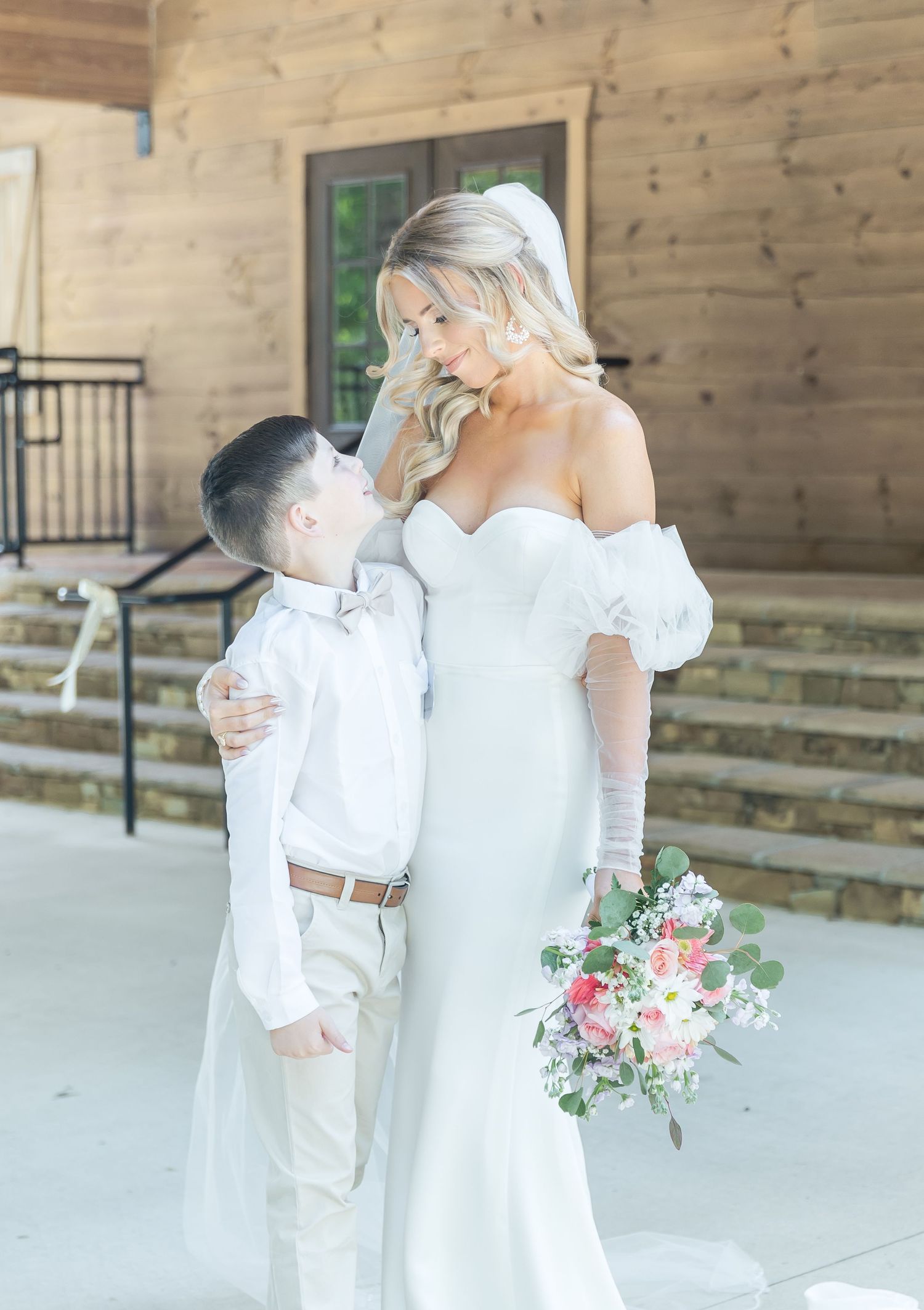 Bride in white off-shoulder gown shares tender moment with young ring bearer on wedding day.