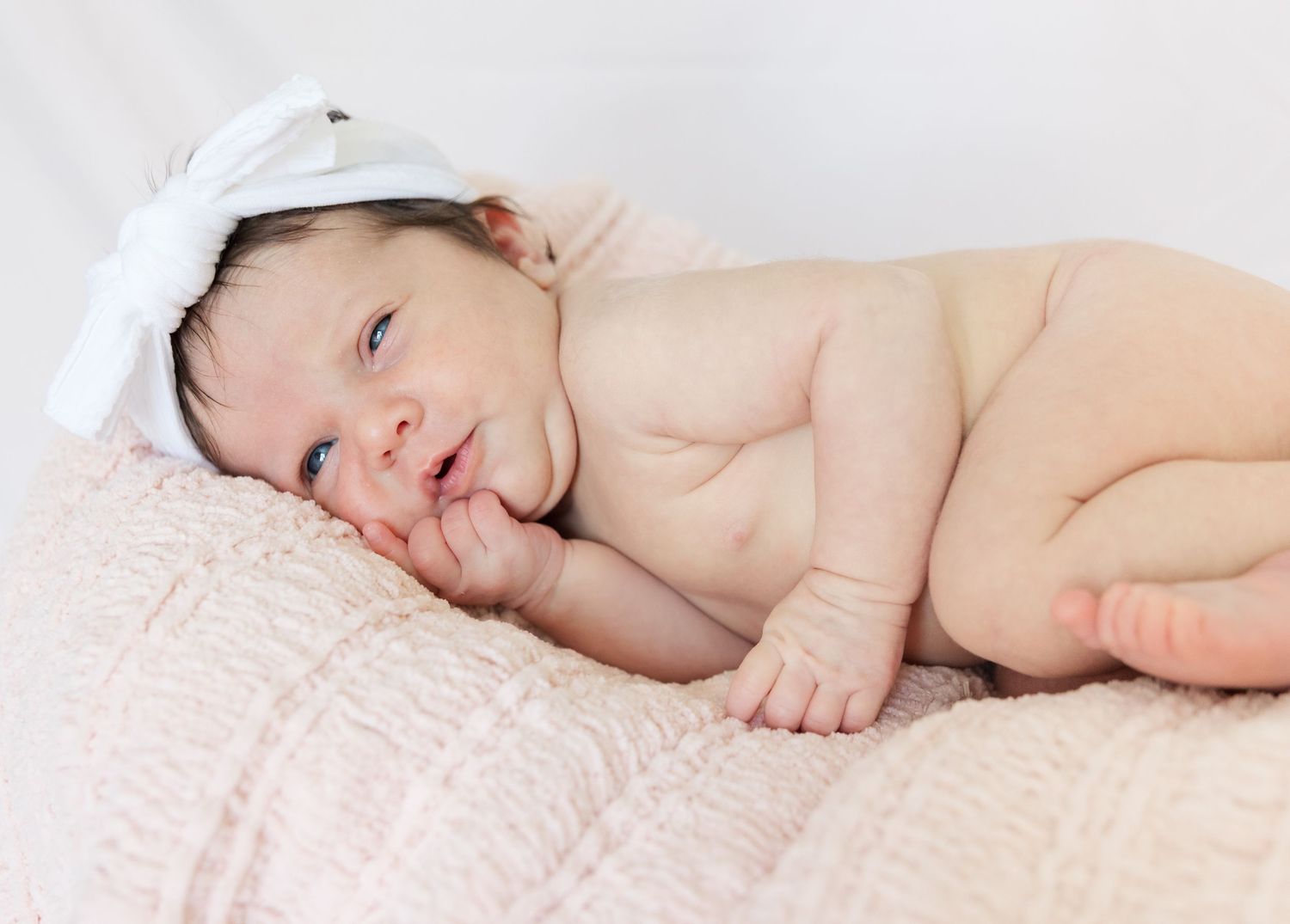 A newborn baby lies on pink bedding wearing a white head covering.