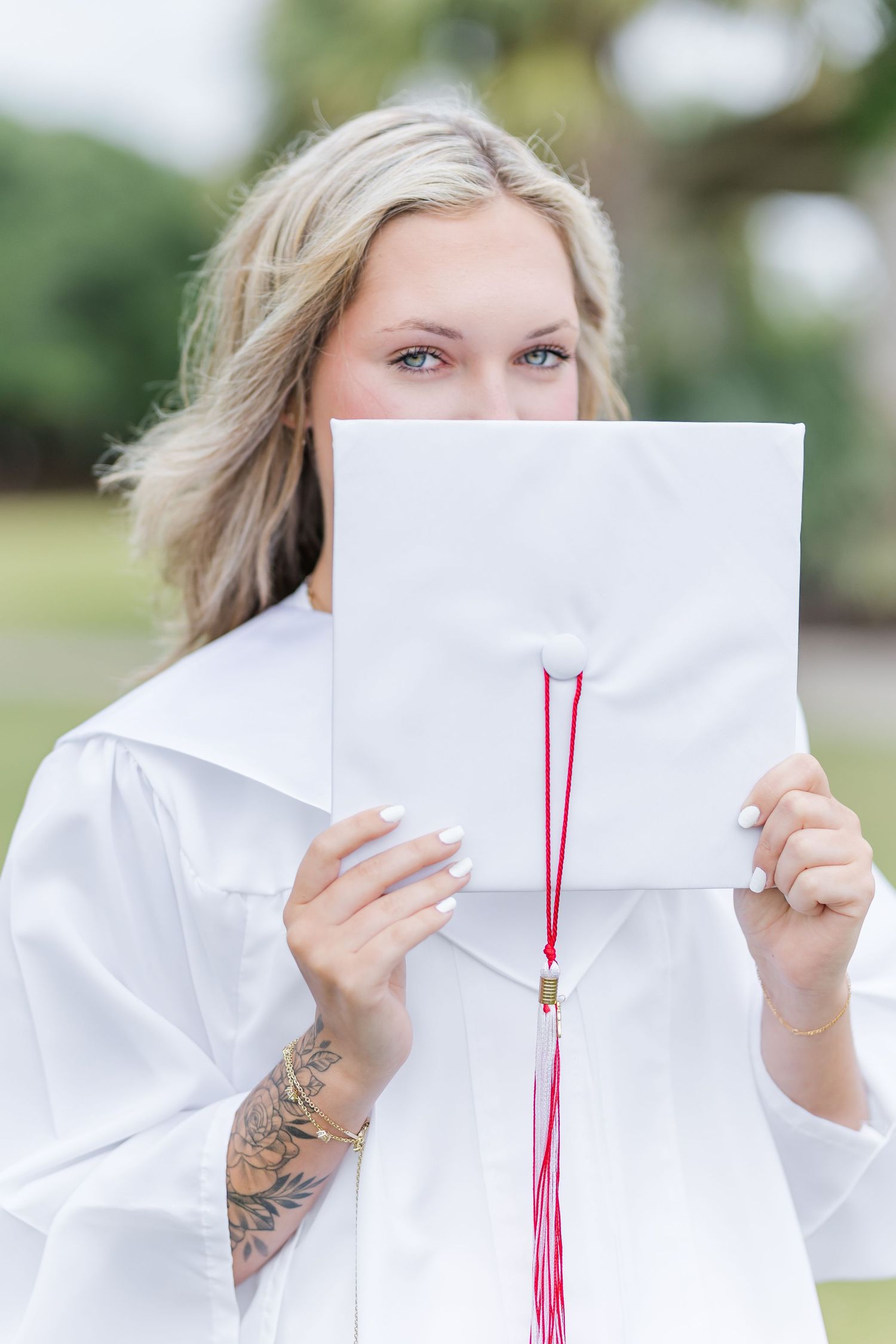 Graduate in white gown holding diploma with red tassel and showing visible arm tattoo.