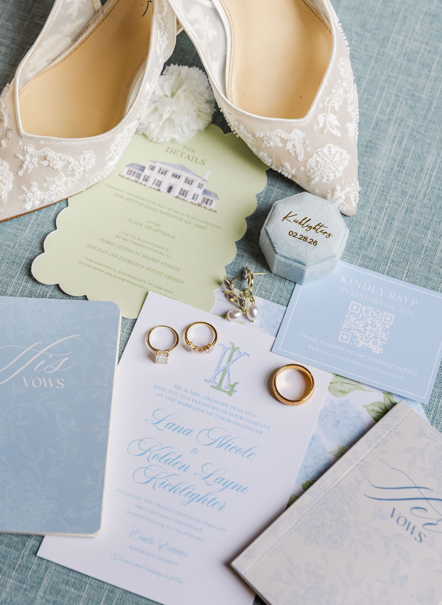 Elegant wedding flat lay with lace bridal heels, gold rings, blue invitations, and vow books on a soft blue surface.