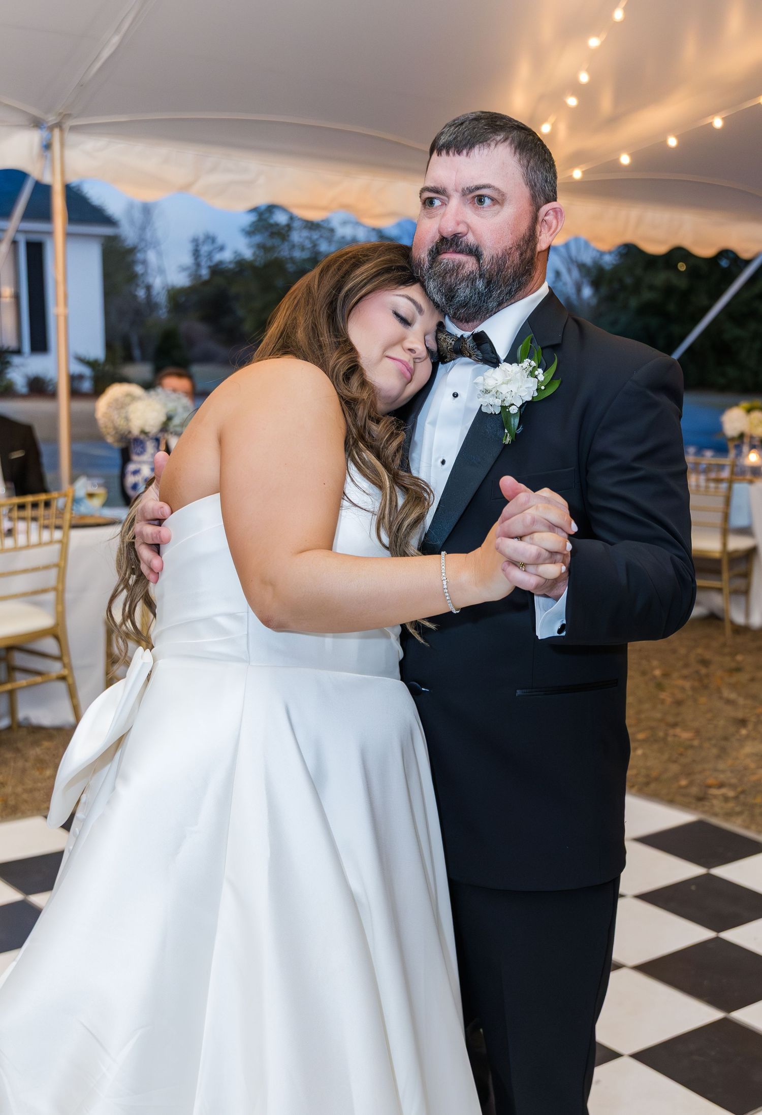 Bride and bearded groom sharing an emotional father-daughter dance under tented reception with string lights.