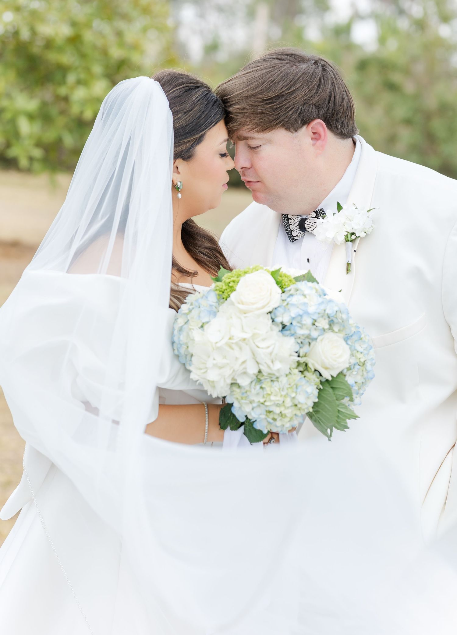 Bride and groom nearly kissing, bride holds blue hydrangea and white rose bouquet, groom wears white tuxedo.
