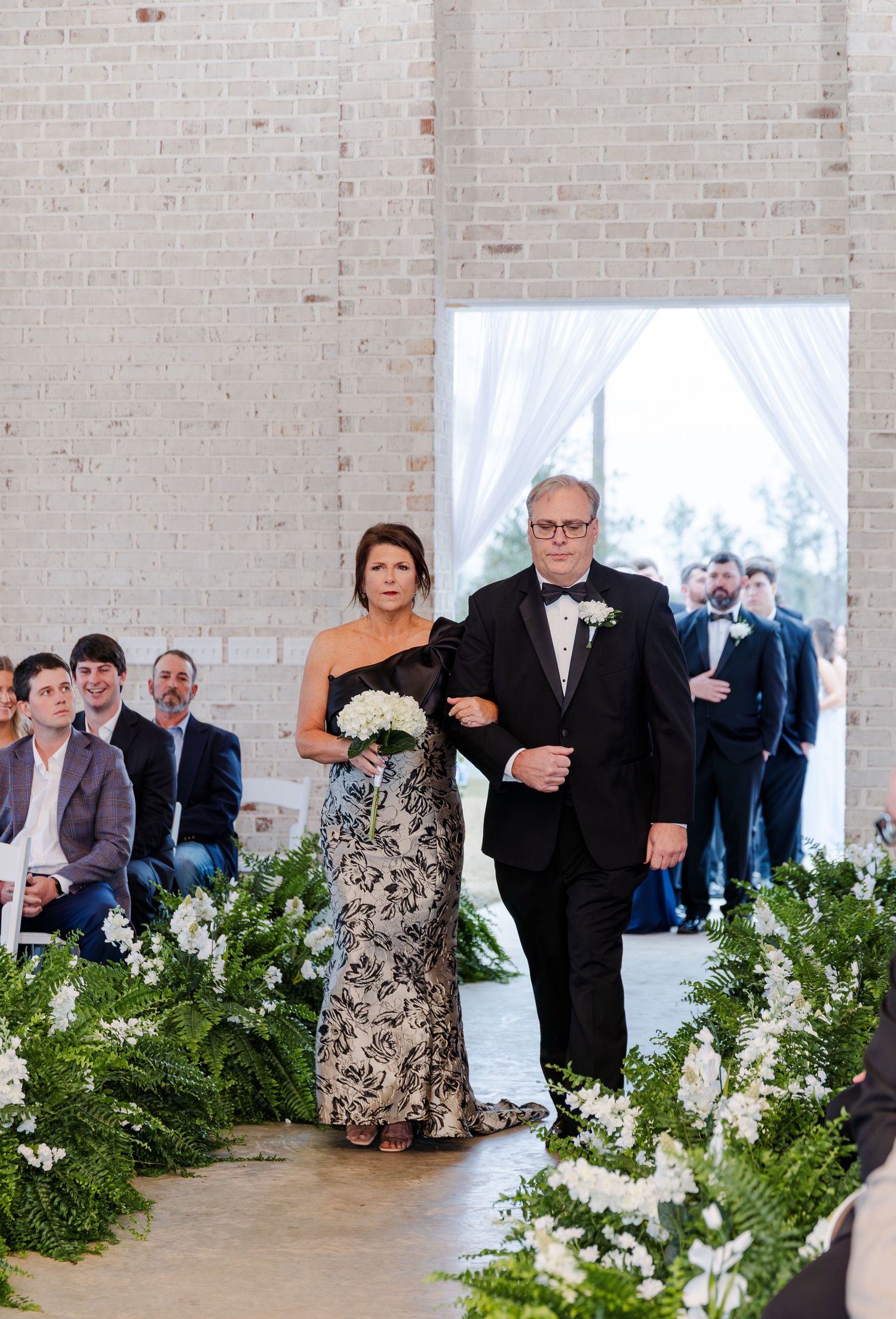 Couple walks down aisle lined with lush greenery in white brick wedding ceremony venue.