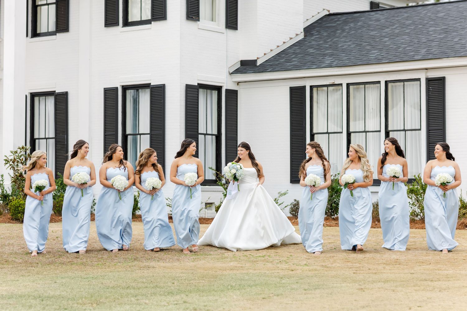 Bride and bridesmaids in light blue gowns walking together outside a white venue with black shutters.