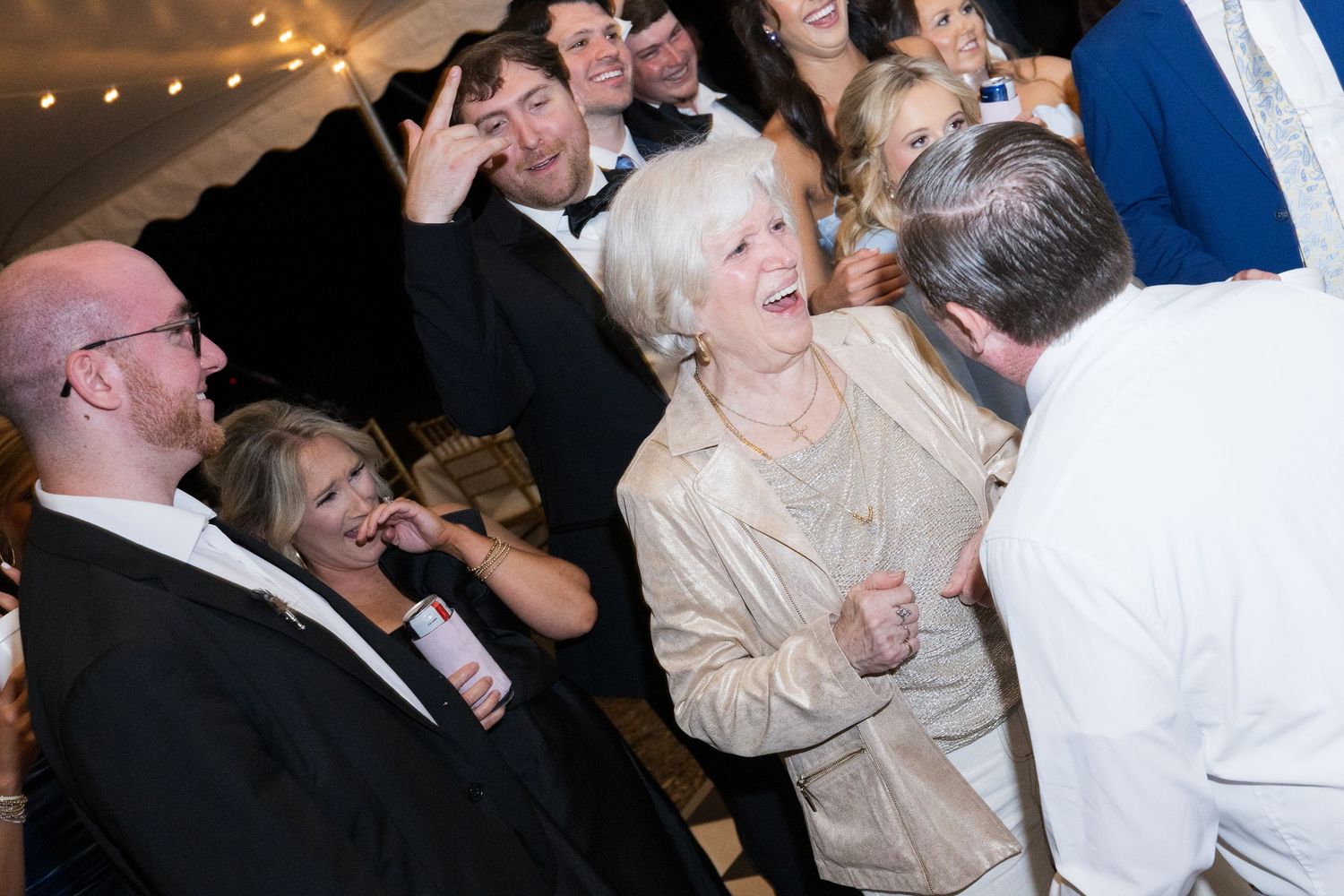 Joyful wedding guests laughing and dancing together under string lights at lively indoor reception celebration.