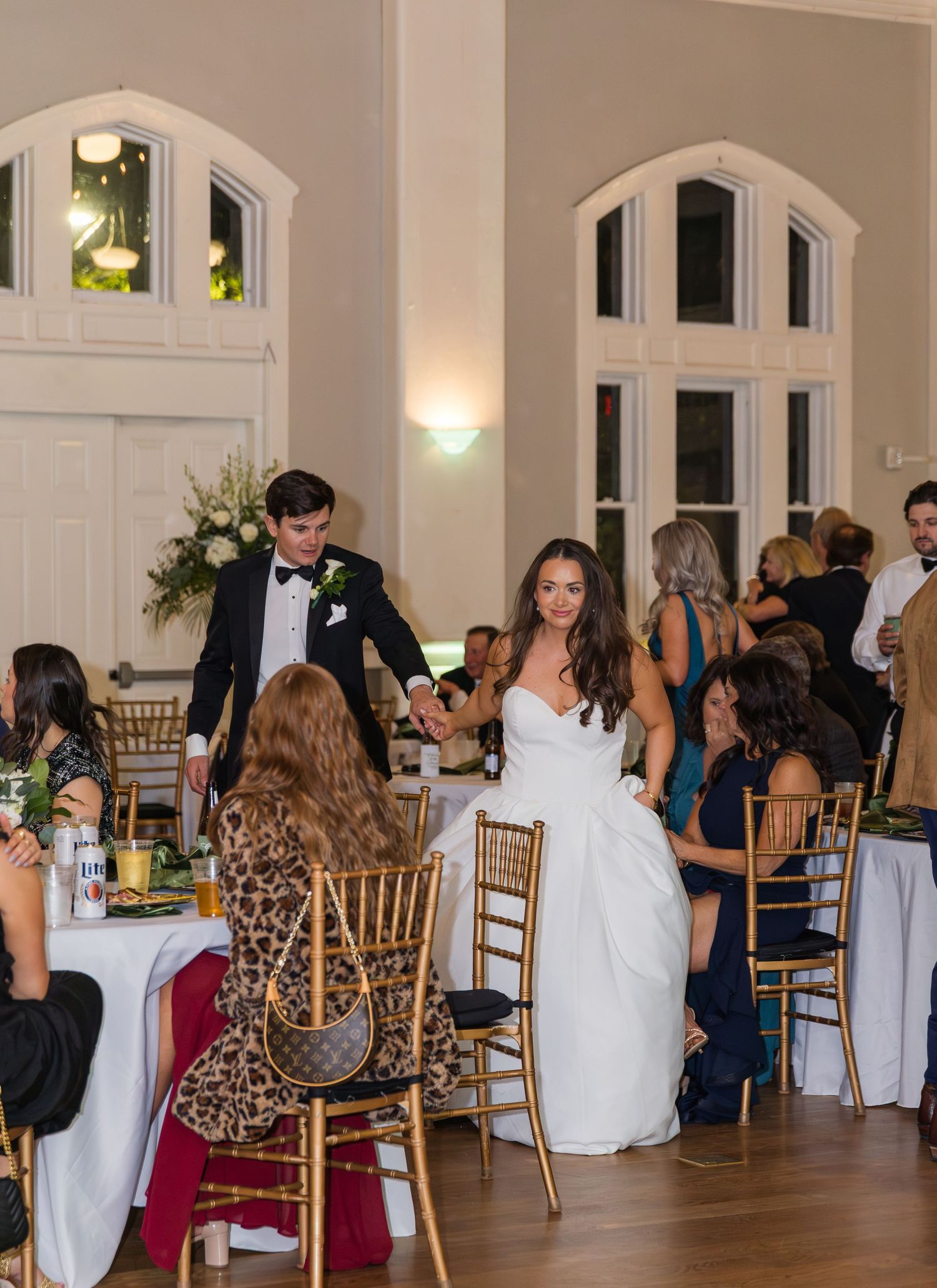 Bride and groom greet wedding reception guests at elegantly decorated venue with gold chiavari chairs.