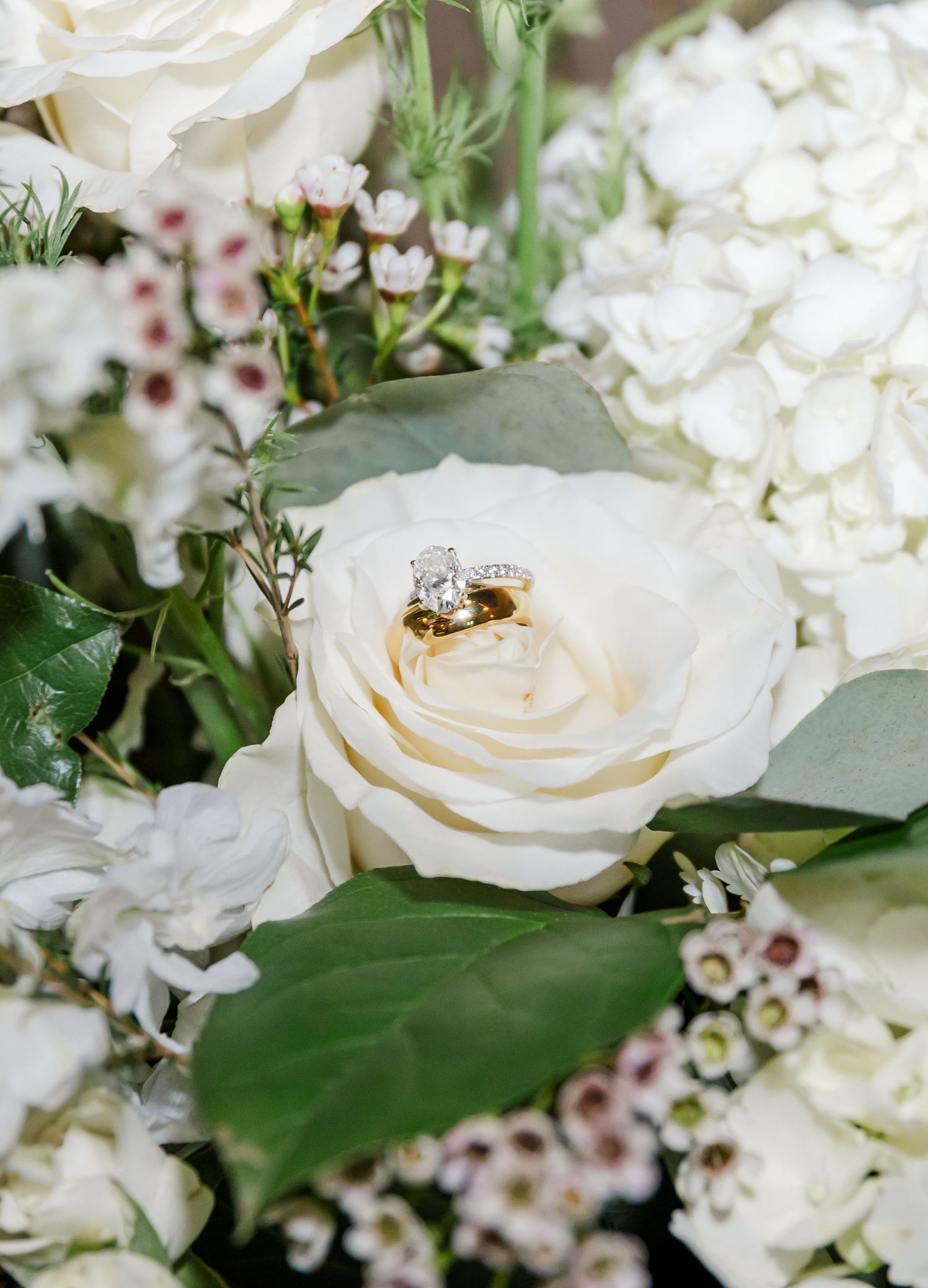 Gold diamond engagement ring and wedding band nestled in white rose surrounded by hydrangeas and greenery bouquet.