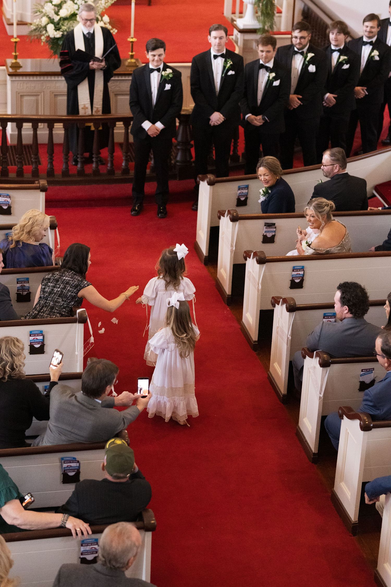 Flower girl walks down red carpet aisle in church wedding ceremony with groomsmen standing at altar.